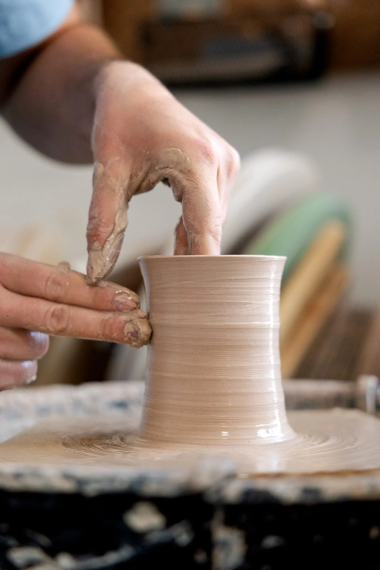 Two wet hands pulling up the wall of a clay cylinder on a pottery wheel in a studio