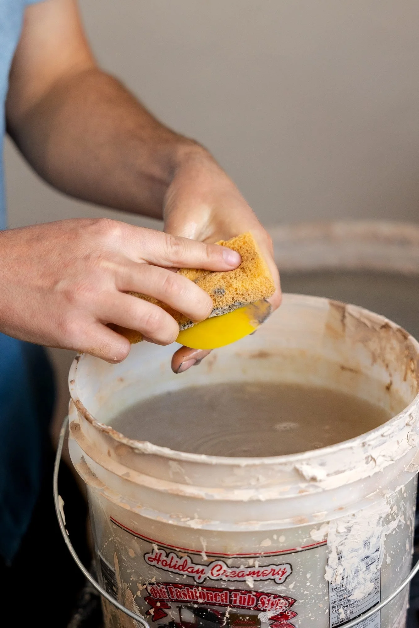 A hand squeezing a yellow sponge over a white bucket filled with wet clay slurry