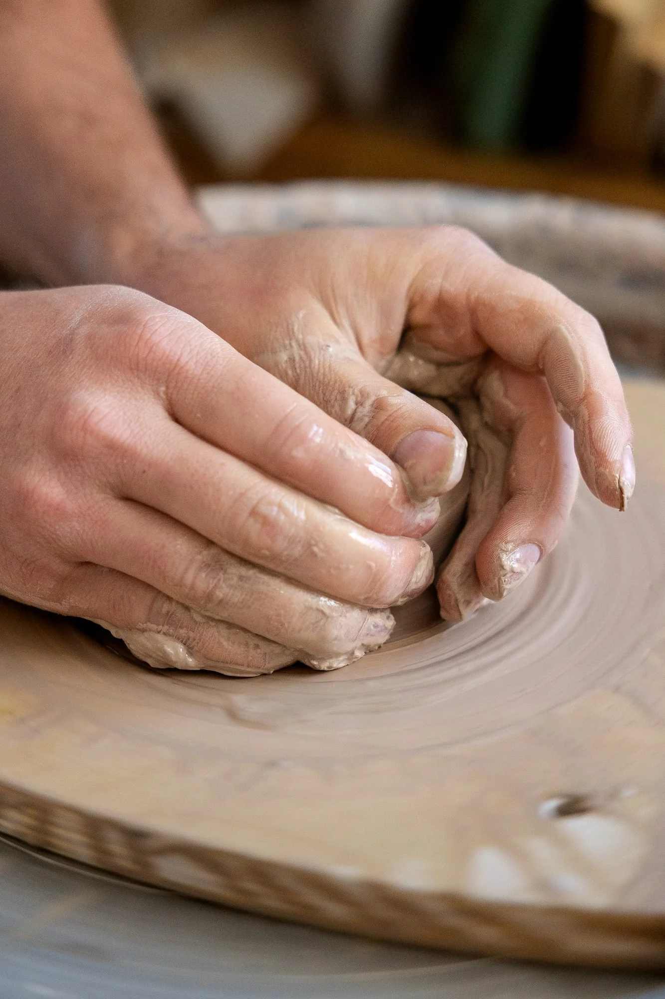 Two wet hands centering a ball of clay on a pottery wheel with a splash pan behind
