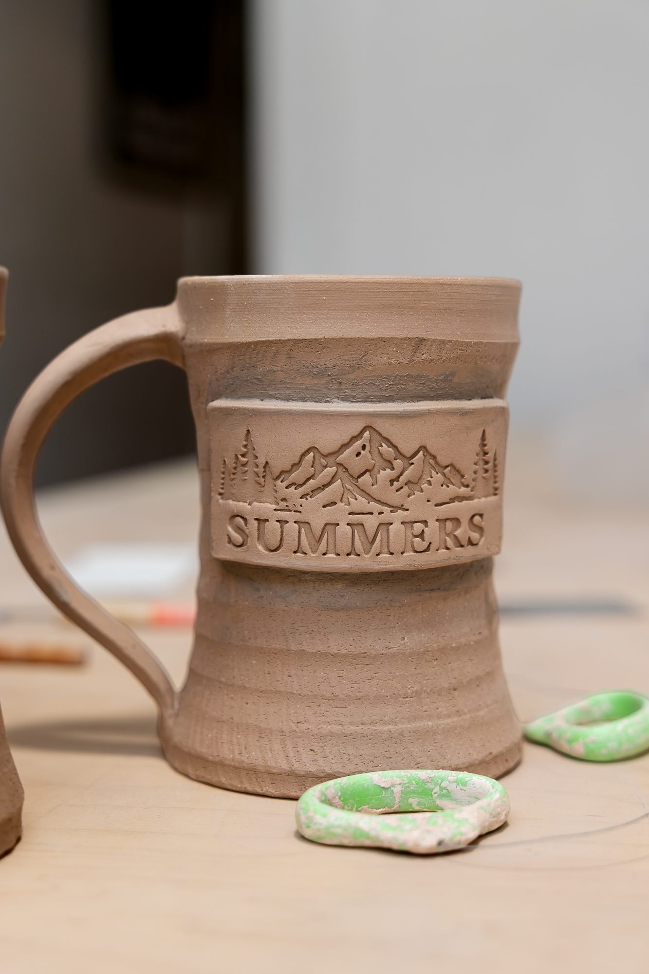A leather hard unglazed clay mug with a pulled handle and a stamped Summers mountain logo on a wooden table