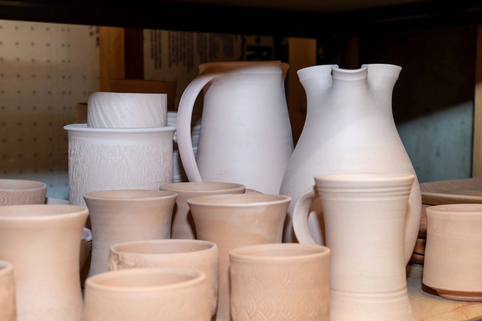 A studio shelf crowded with pale pink bisque fired pots including a pitcher, bowls, and cups