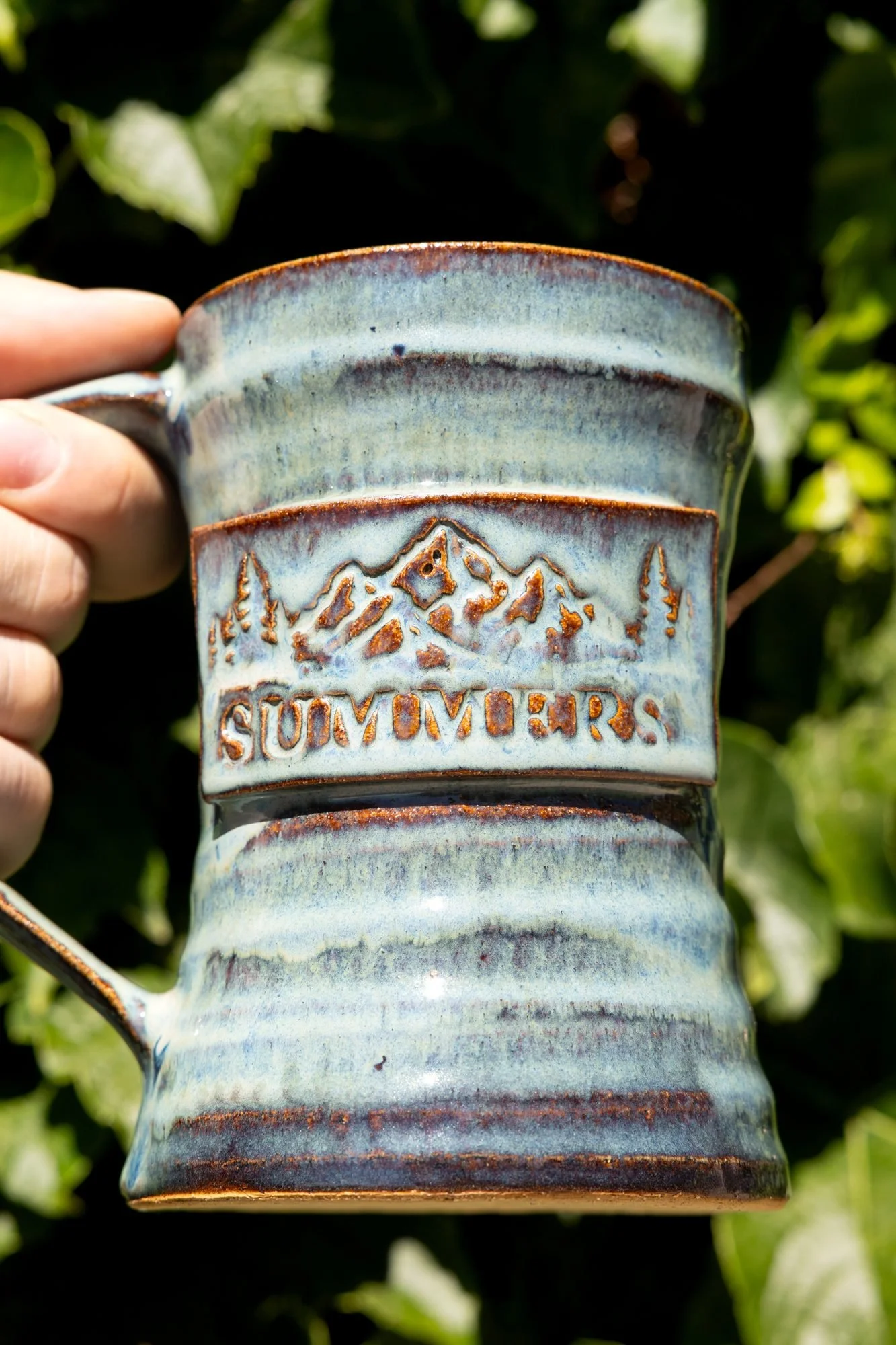 A hand holding a blue glazed mug with a stamped Summers logo against a wall of green ivy