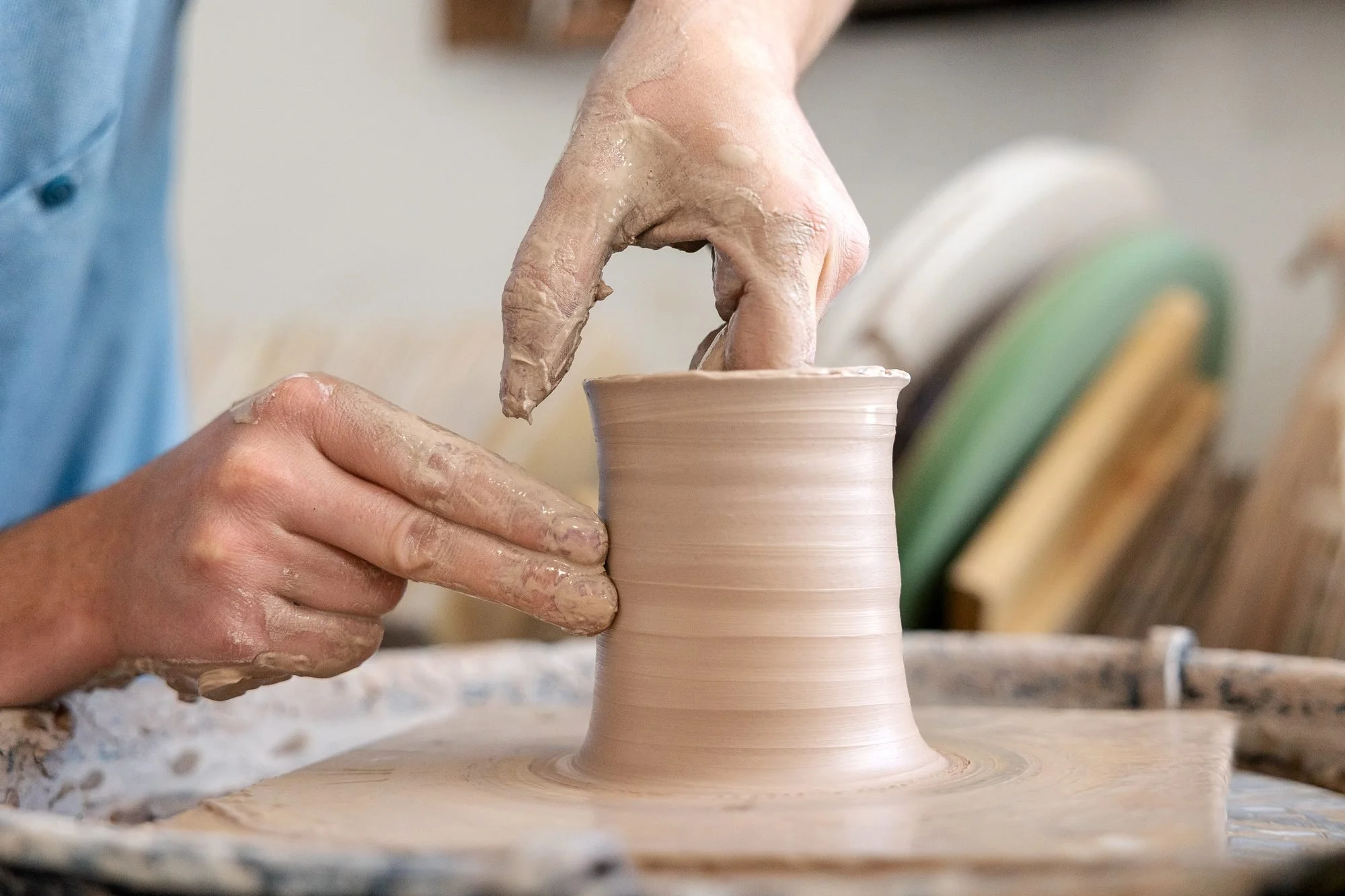 Two wet hands pulling up the wall of a clay cylinder on a pottery wheel with tools on the splash pan