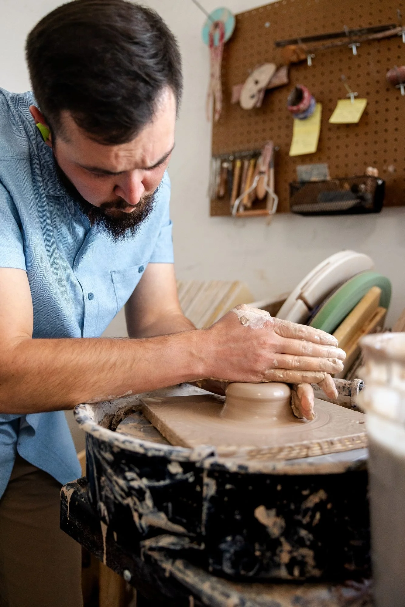 Adam Corbridge in a blue shirt seated at the wheel, both hands wrapped around a lump of clay as he centers it