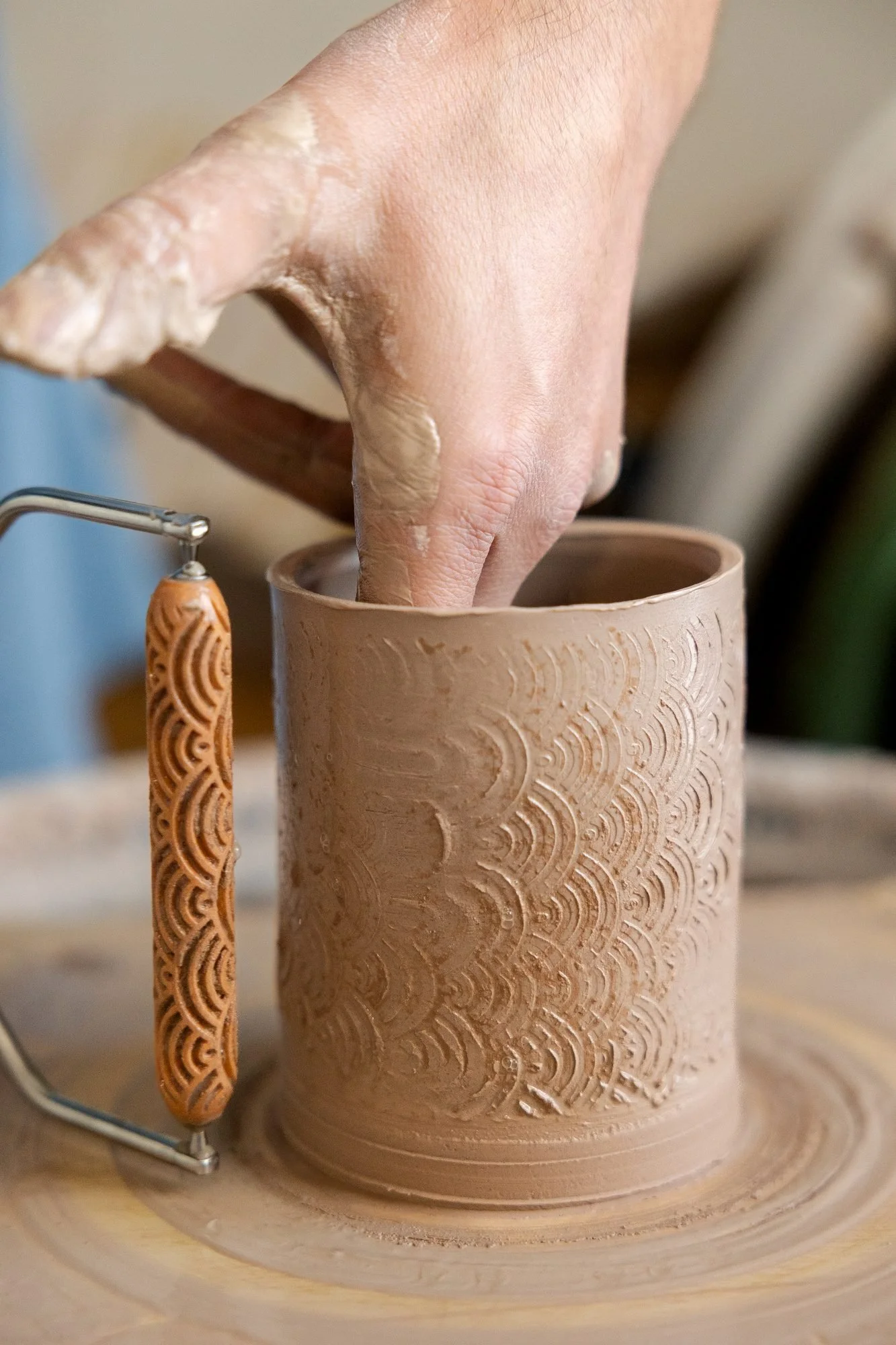 A wire handled texture roller being pressed against the side of a wet clay cylinder while a hand supports it from inside