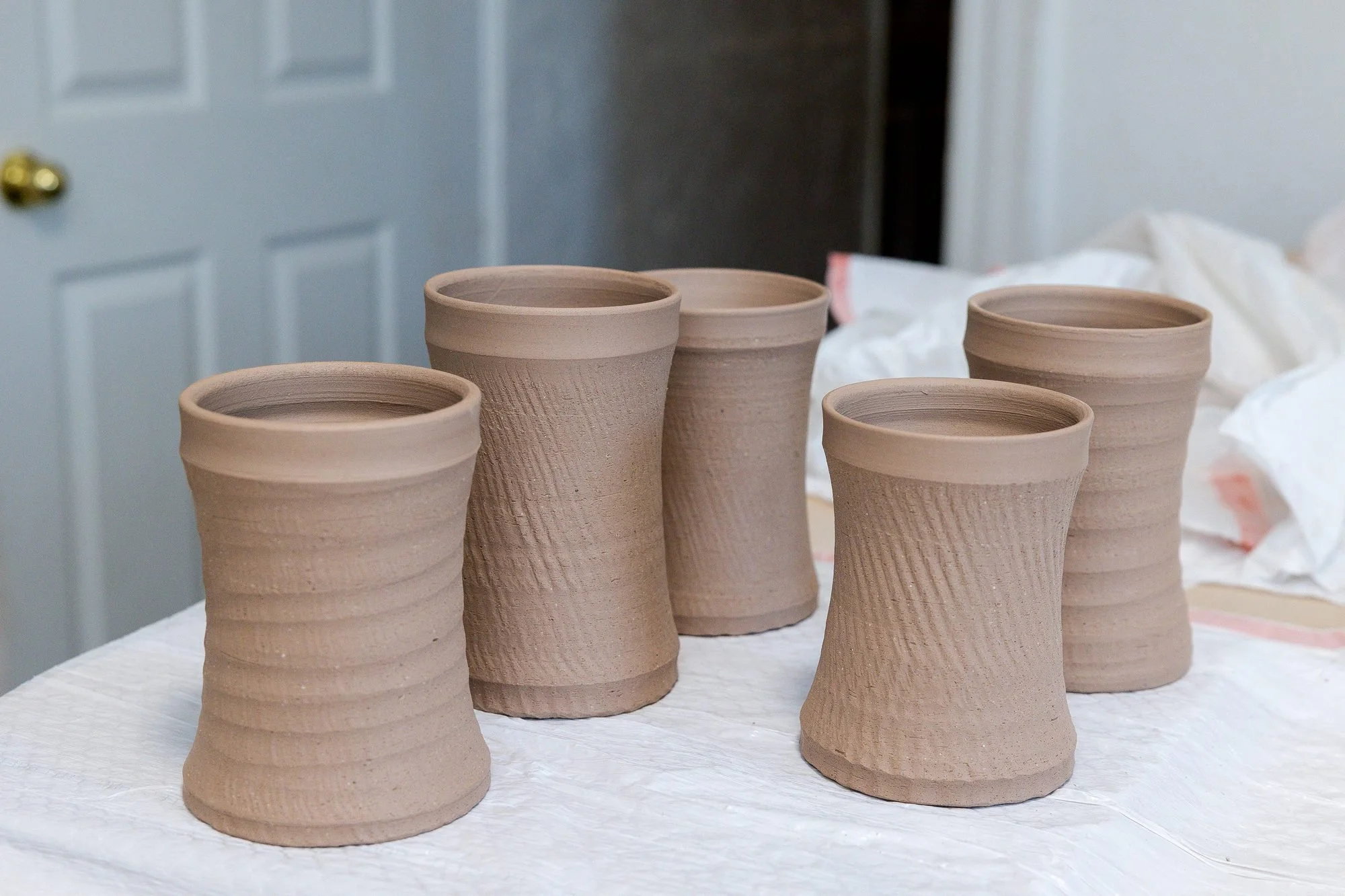 A group of freshly thrown unglazed clay mug bodies with flared rims standing on a cloth covered table near a white door
