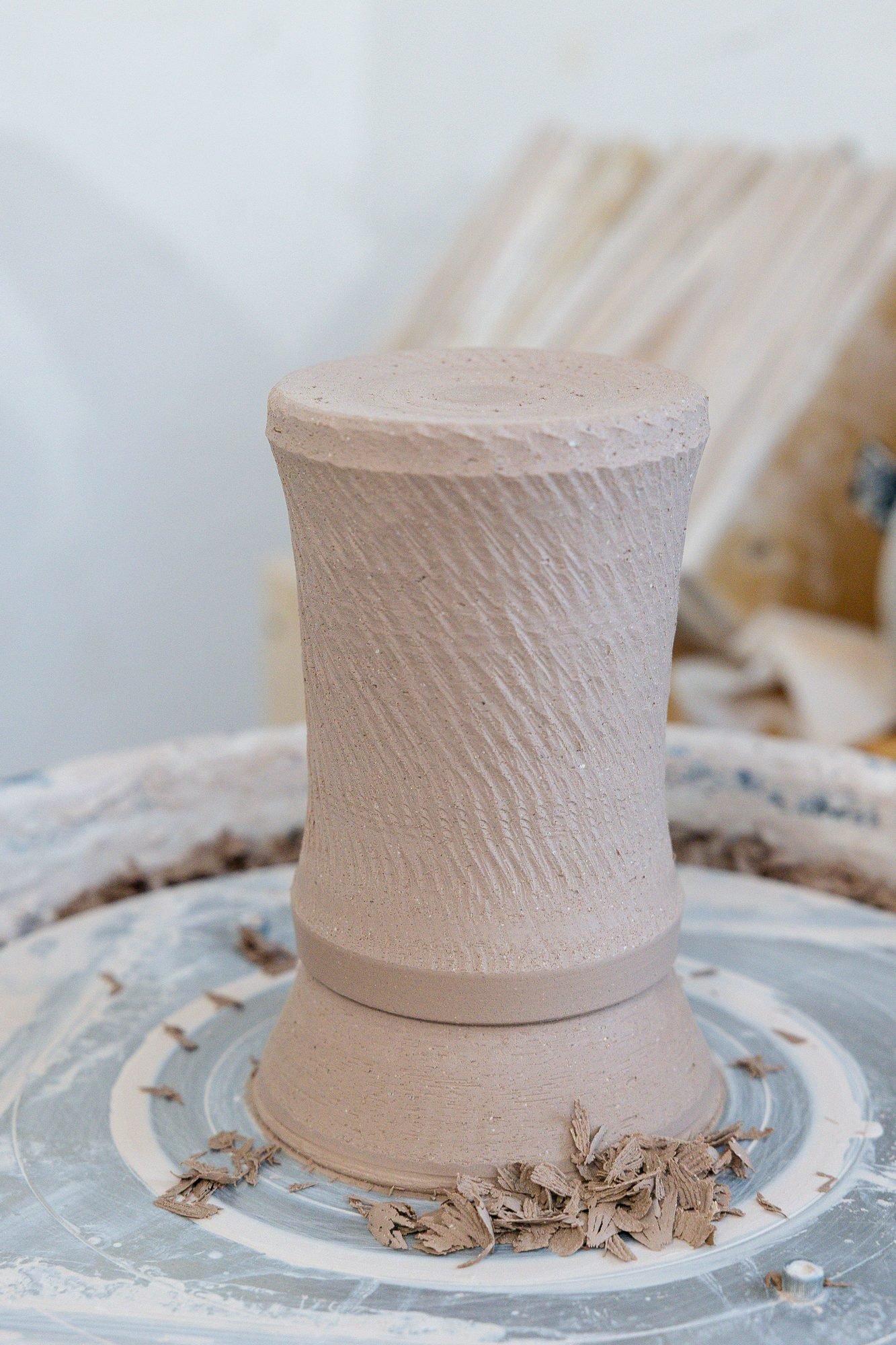 A textured clay mug sitting upside down on a pottery wheel surrounded by curled clay trimmings after its foot was trimmed