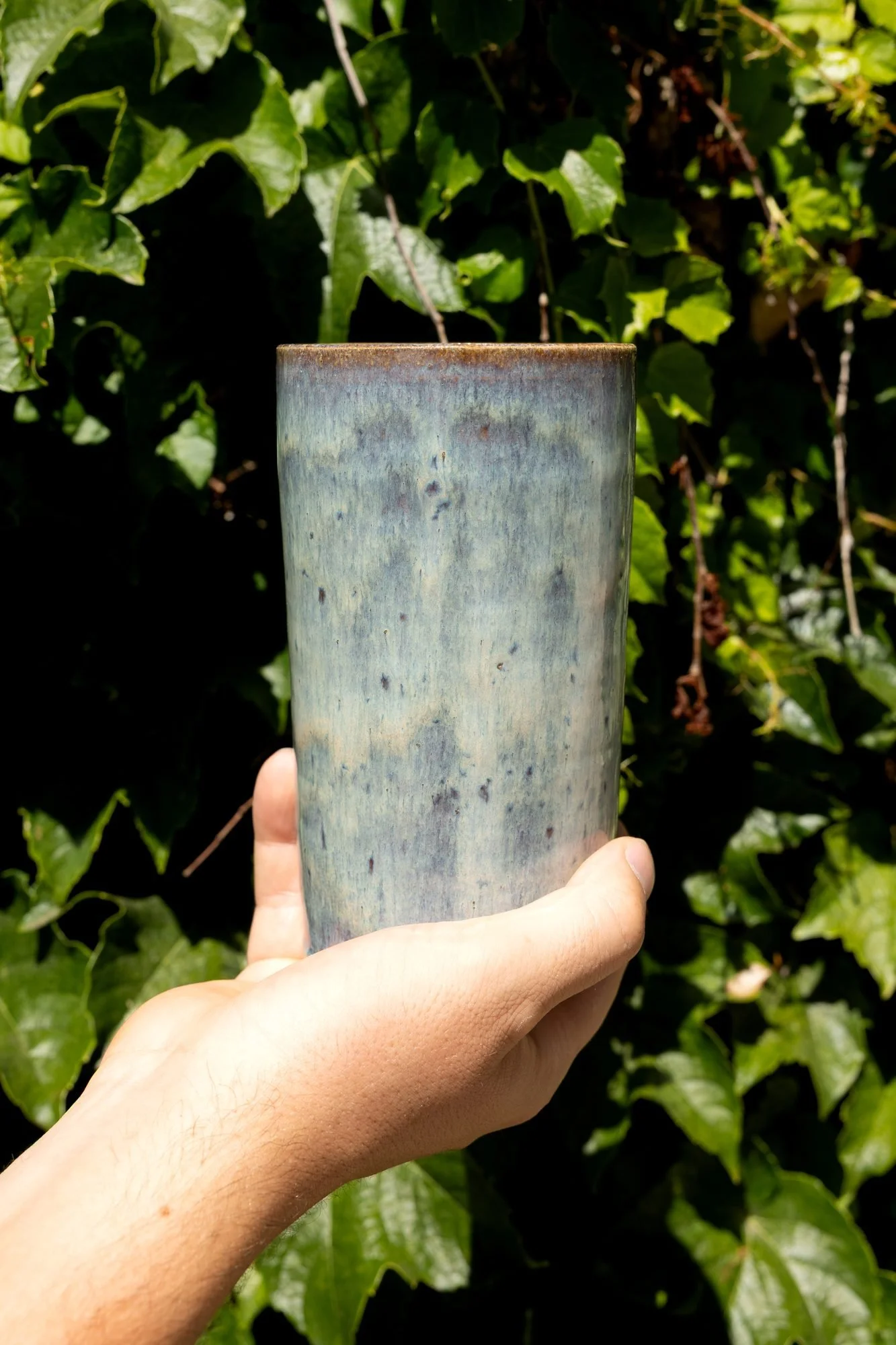 A hand holding a tall blue glazed tumbler against a background of green ivy leaves