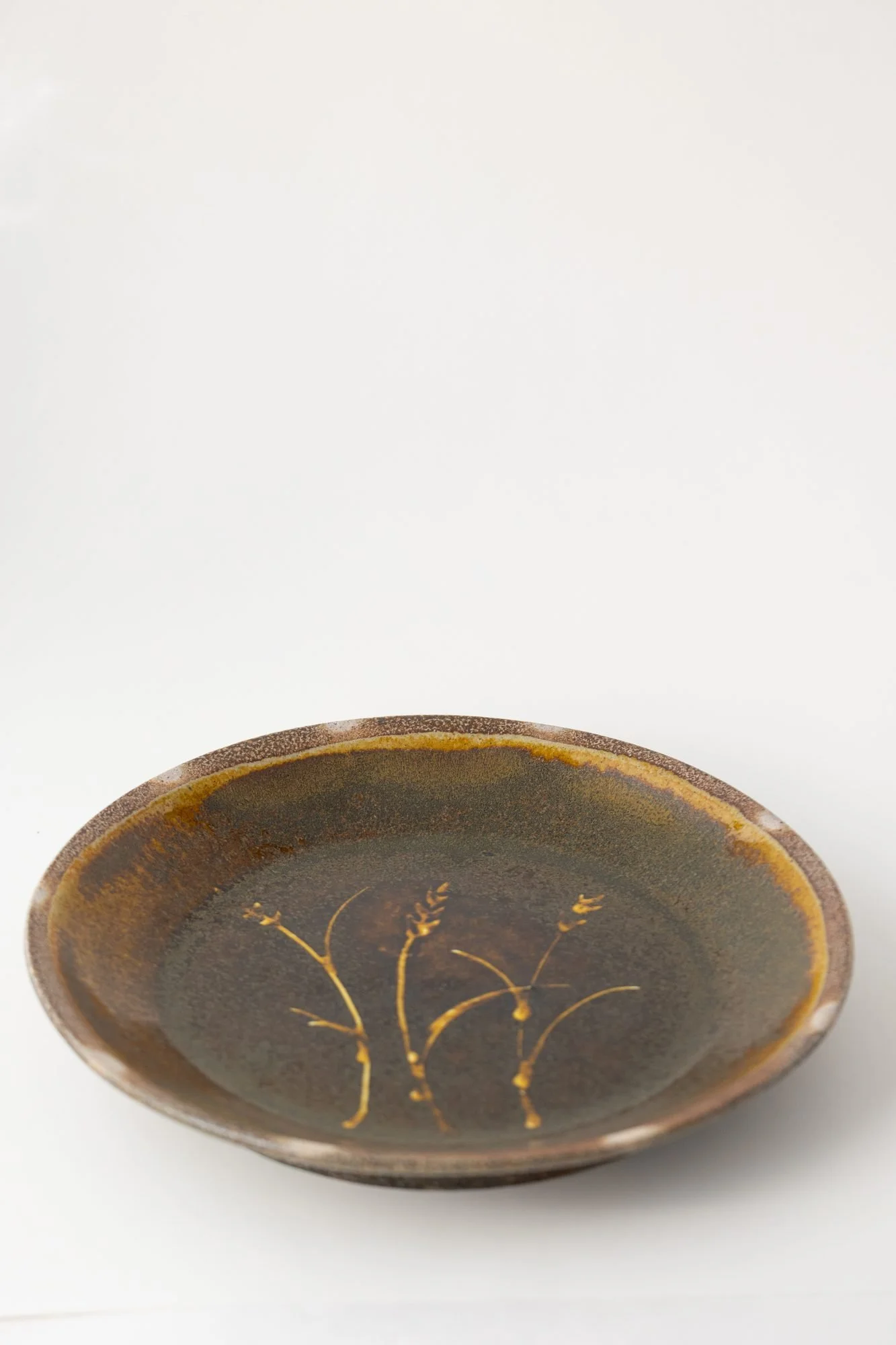 A small brown glazed plate with a painted design of wheat stalks in the center on a white background