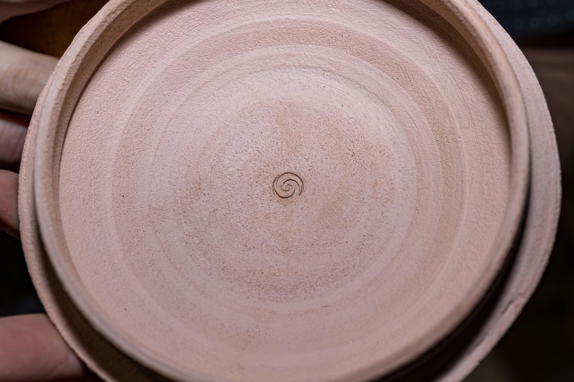 Hand holding a trimmed bisque pot base upward, showing a small spiral mark pressed in the center