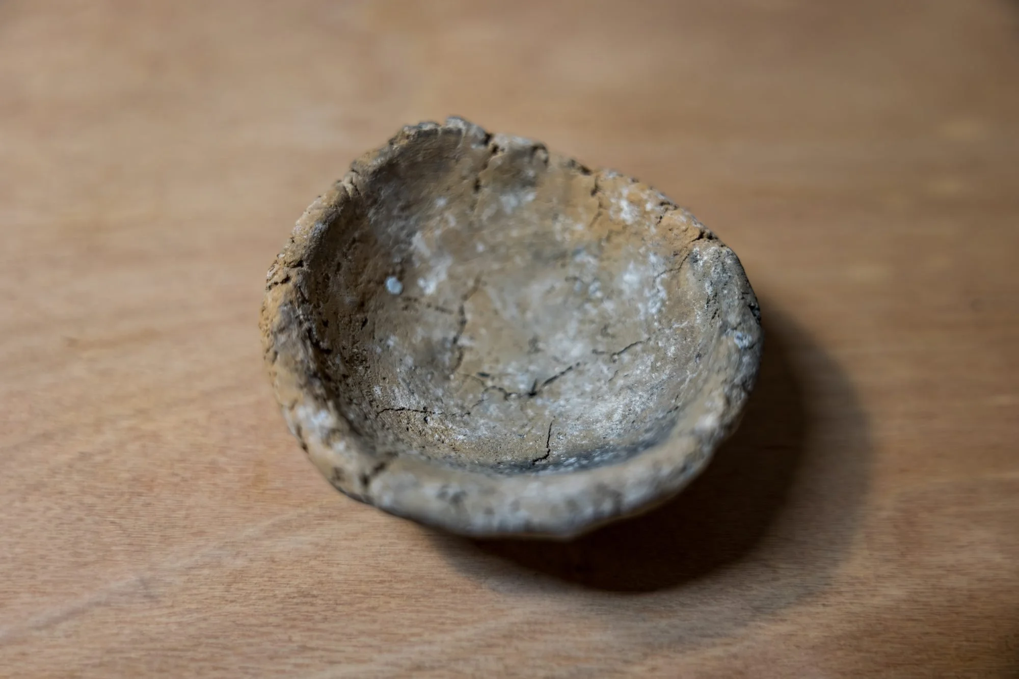 A small rough pinched bowl of gritty grey wild clay on a wooden table, edges uneven and cracked