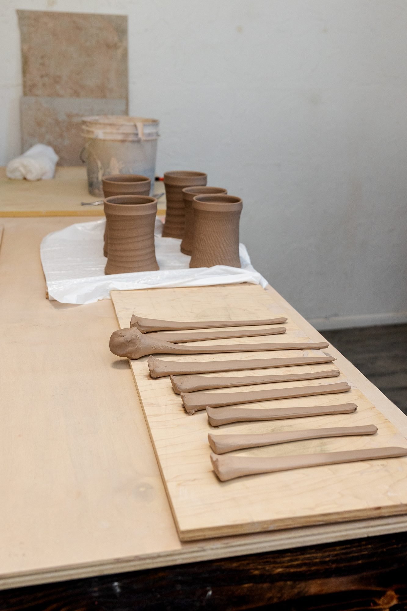 A row of pulled clay handles laid out on a plywood board with thrown mugs waiting on a cloth behind them