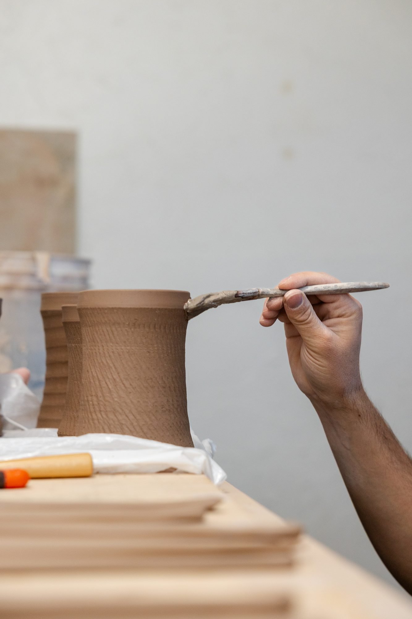 A hand using a small pointed tool to score the side of a textured unfired clay mug near the rim