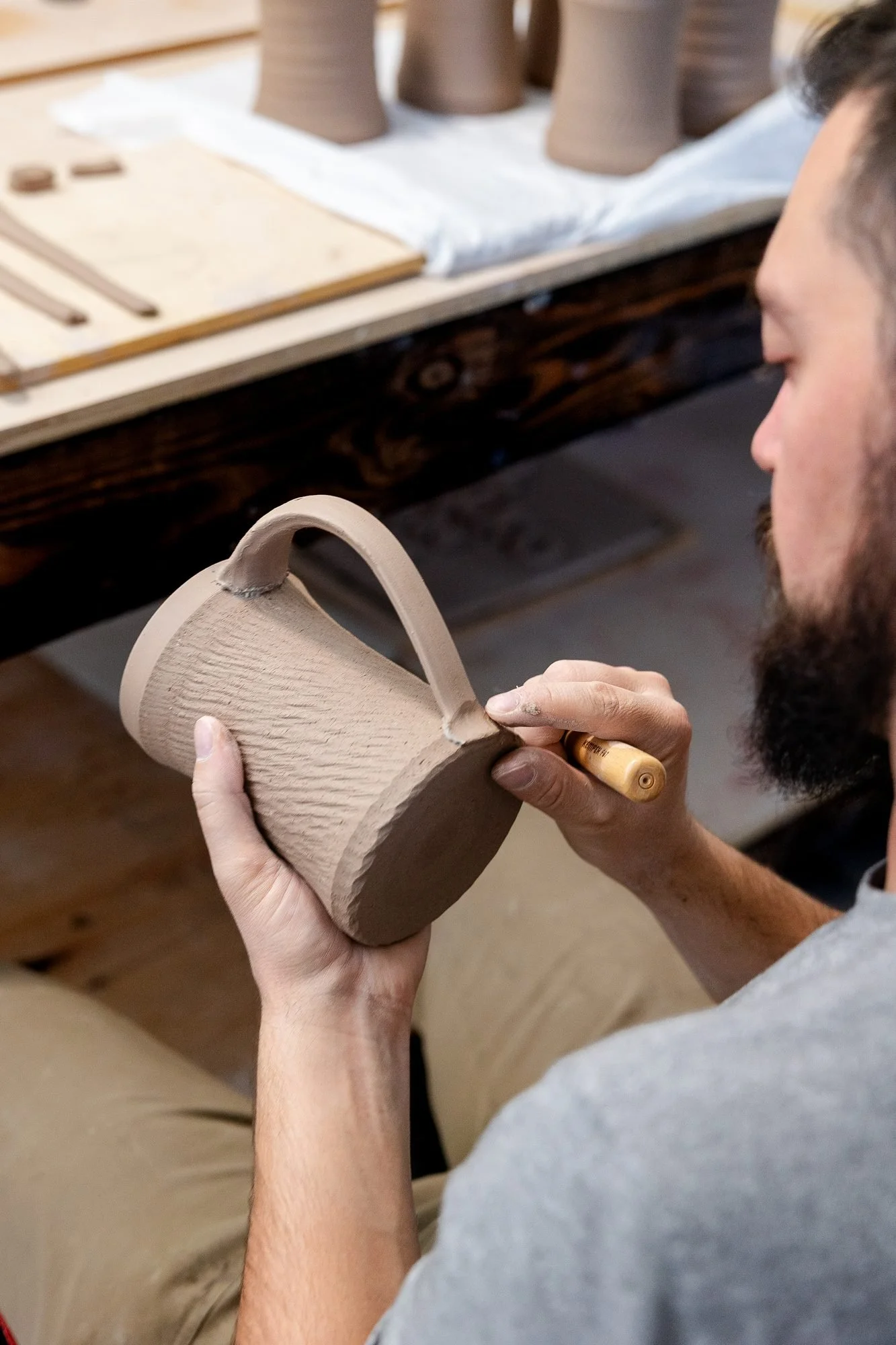 A hand holding a textured mug with a fresh handle while a wooden tool smooths the lower handle join