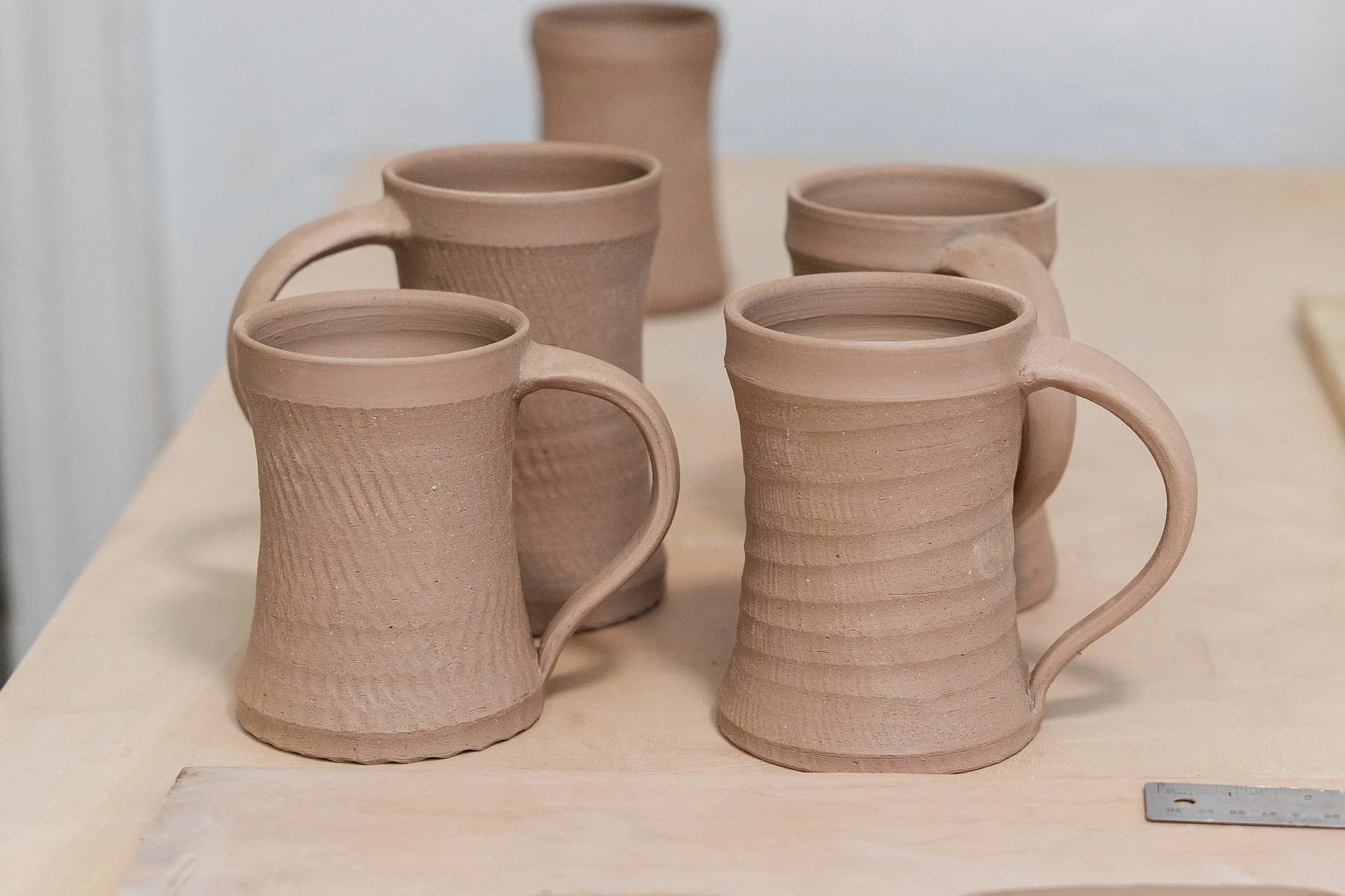 Five handled unfired stoneware mugs standing on a work table with a stamped nameplate slab beside them