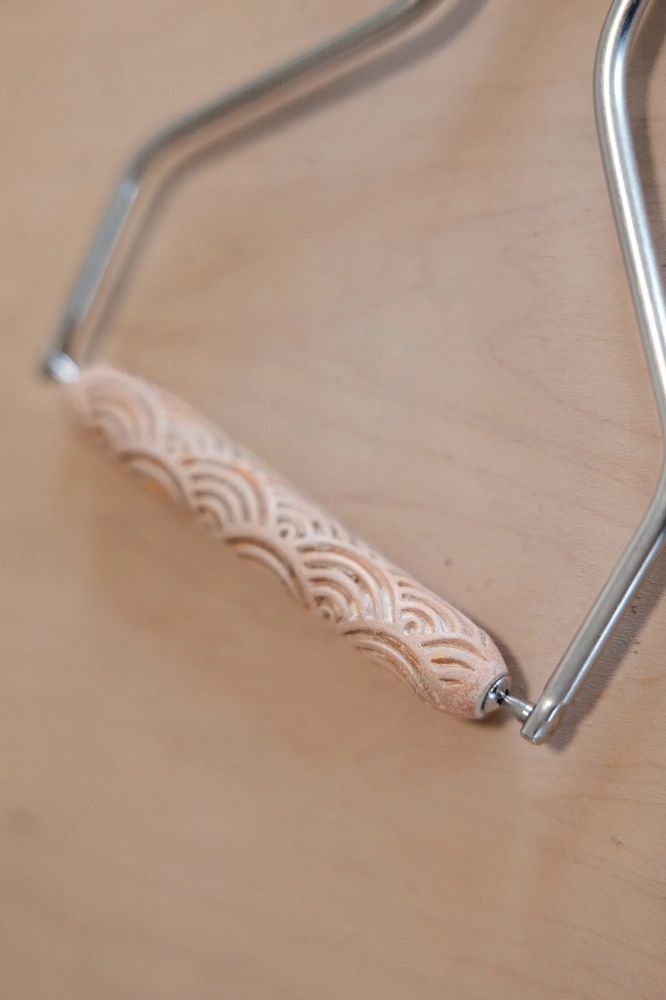 A textured clay roller with a scalloped pattern mounted on a bent wire handle lying on a wooden board