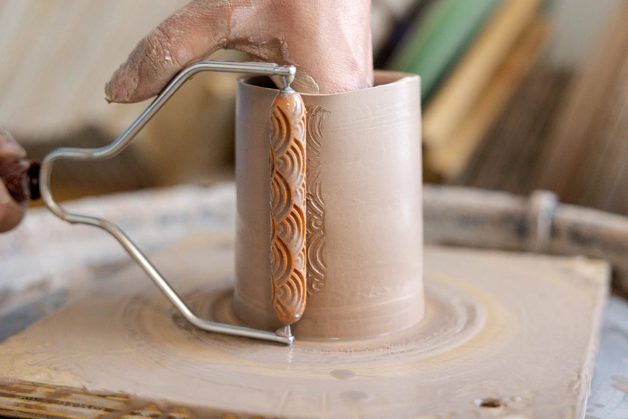 A scalloped texture roller being pressed into the side of a freshly thrown clay mug on a pottery wheel