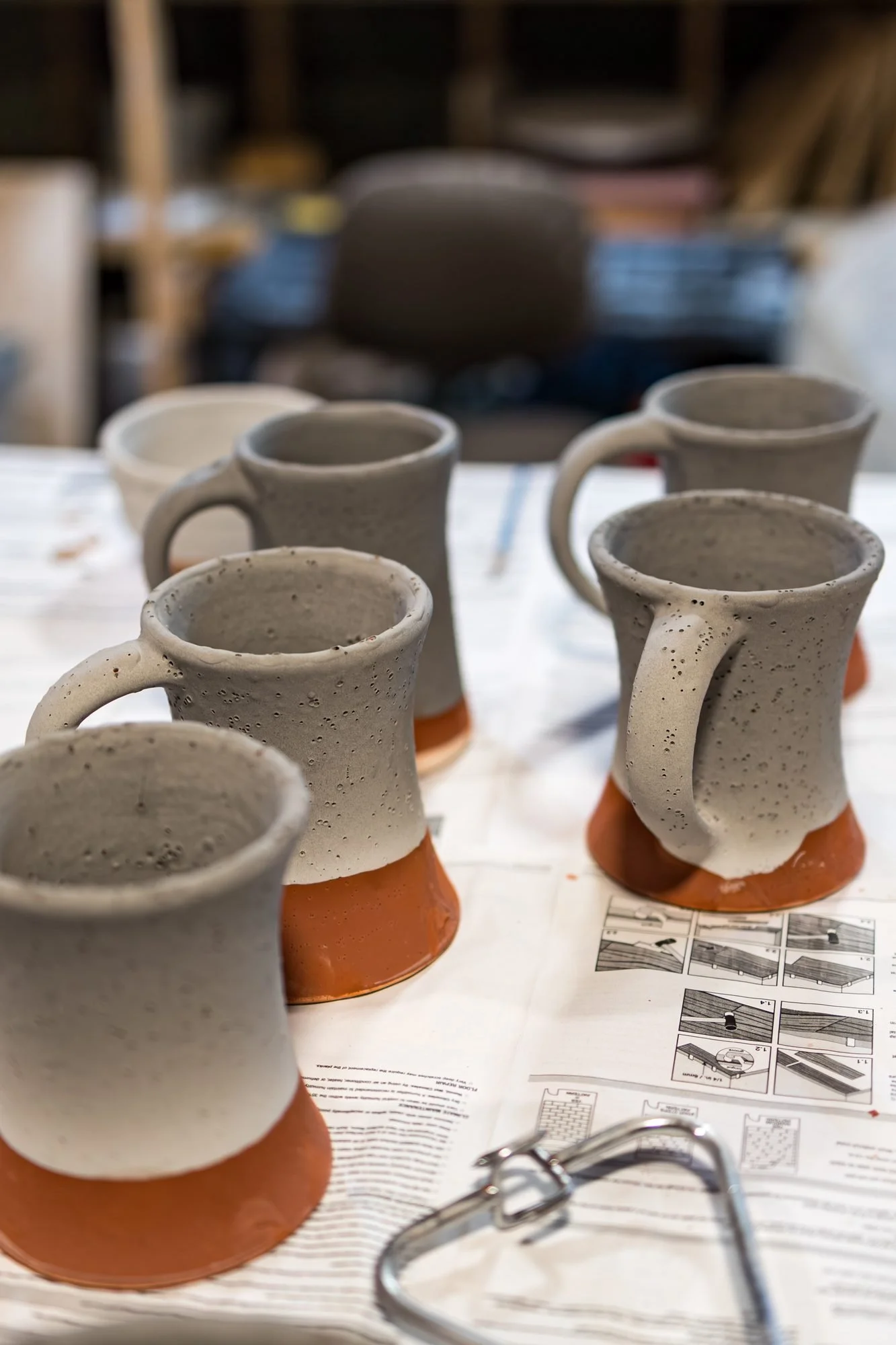 Several mugs coated in matte grey unfired glaze with bare red clay feet standing on newspaper