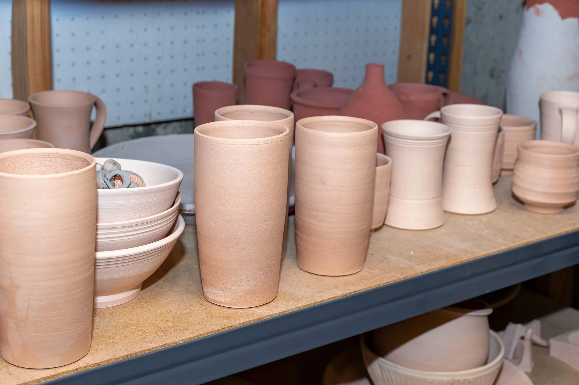 Pale pink bisqued mugs, cups and bowls crowded on a wooden shelf in front of a pegboard wall