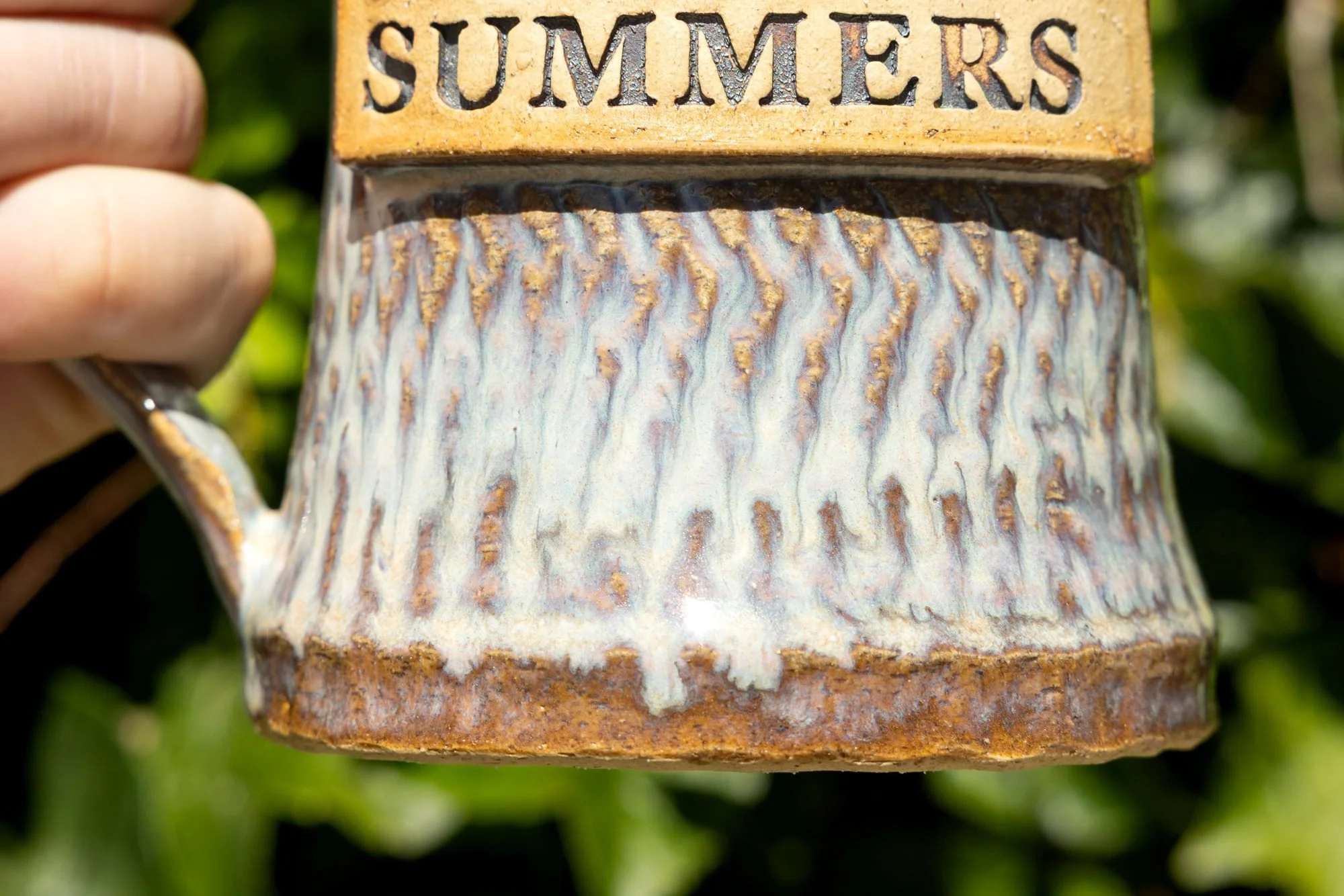 Close view of a finished mug with a stamped SUMMERS panel above textured blue white glaze melted over combed lines