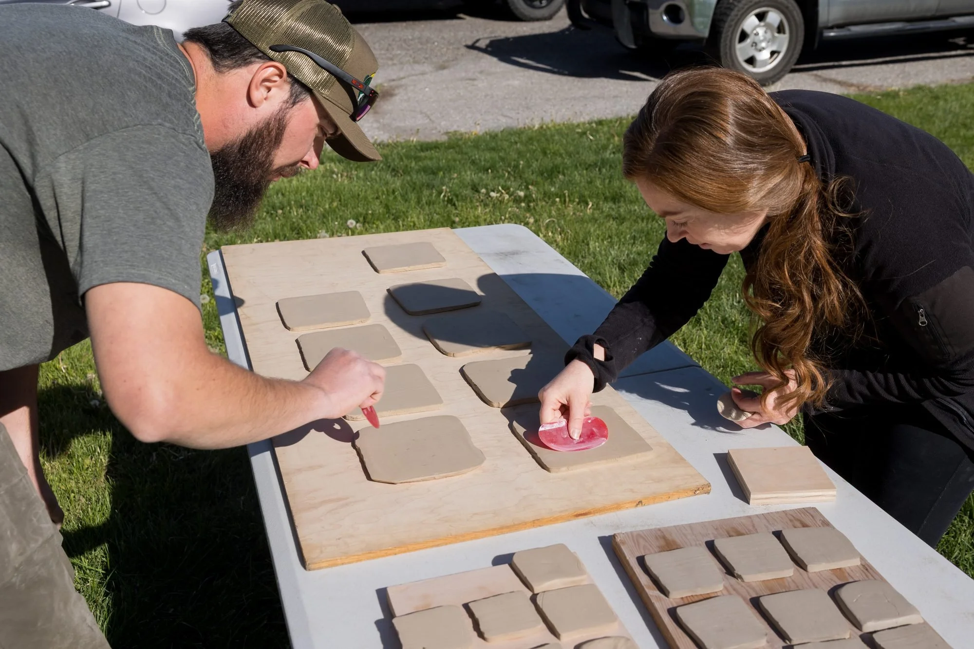 Two people at an outdoor table pressing and arranging clay tiles on boards