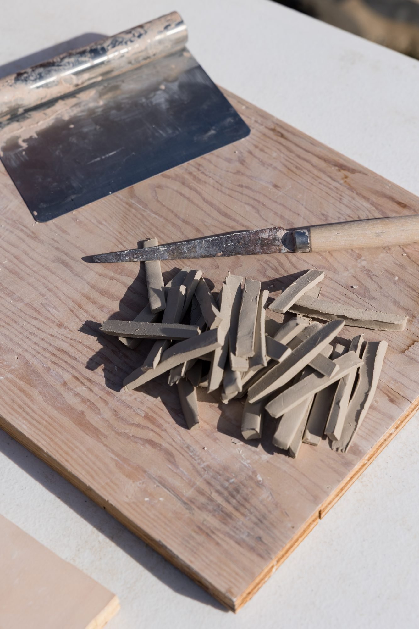 A pile of clay offcuts next to a scraper and a knife on a wooden board