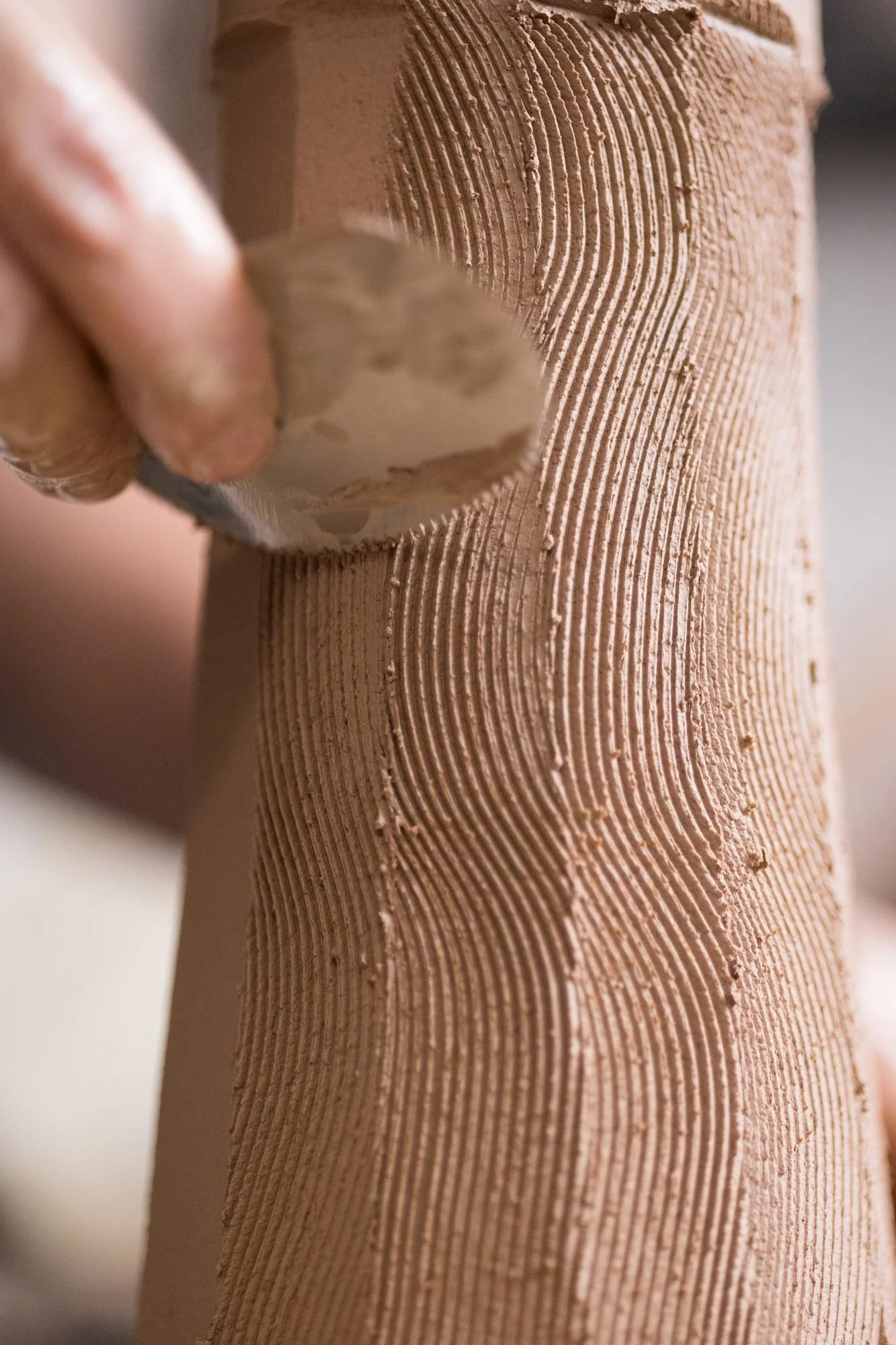 Hands pressing a rib into the wall of a tall clay form, leaving ribbed lines