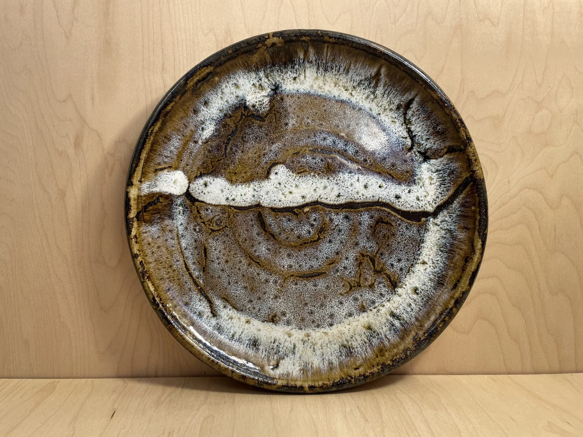 A round stoneware plate with a white and brown glaze and a spiral center, standing on a wooden table