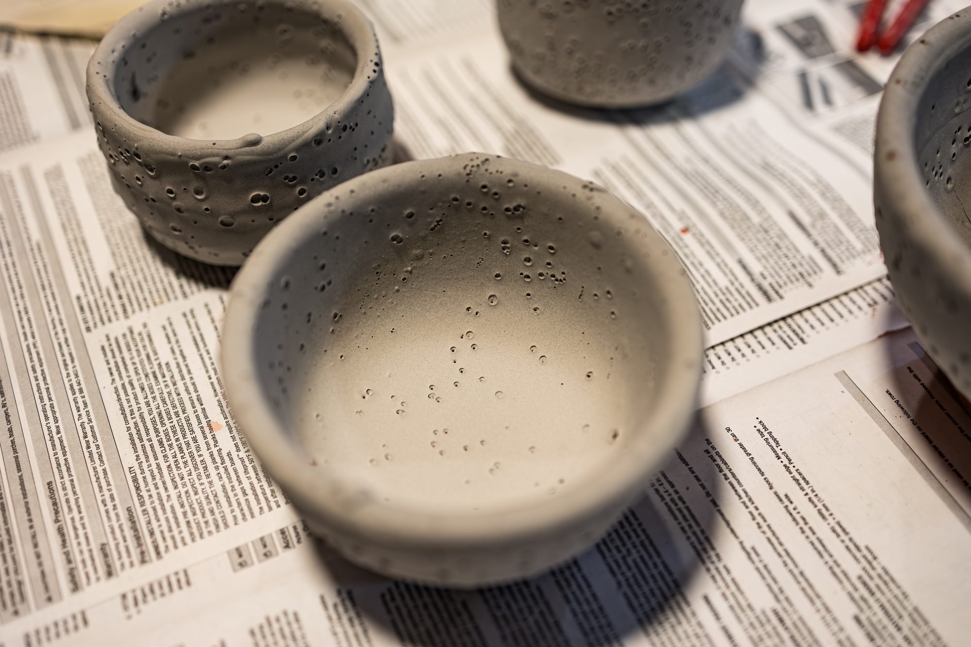 Bisque bowls coated in raw grey glaze with small dark specks, resting on newspaper