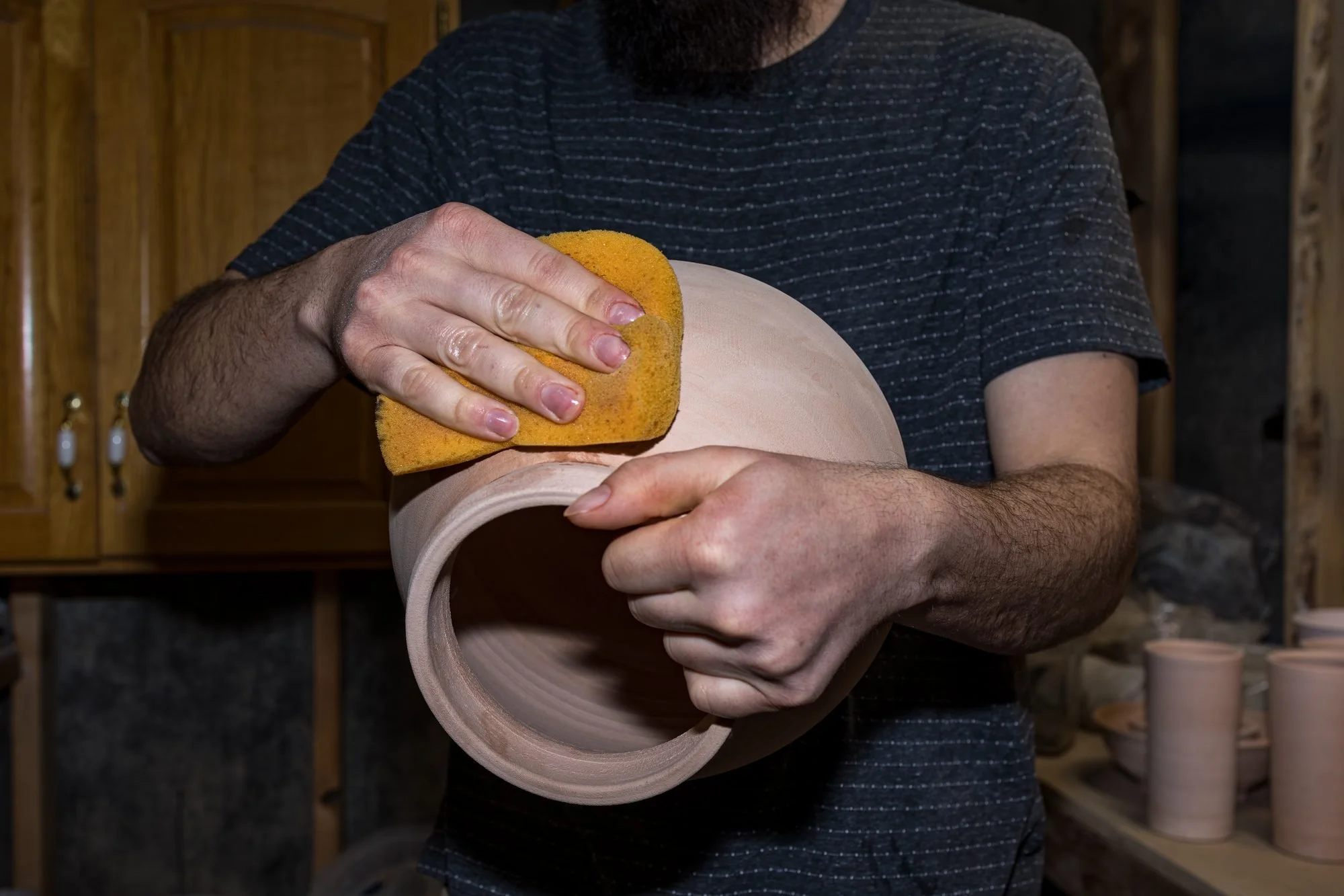 A hand wiping the outside of a bisque fired pot with a yellow sponge before glazing