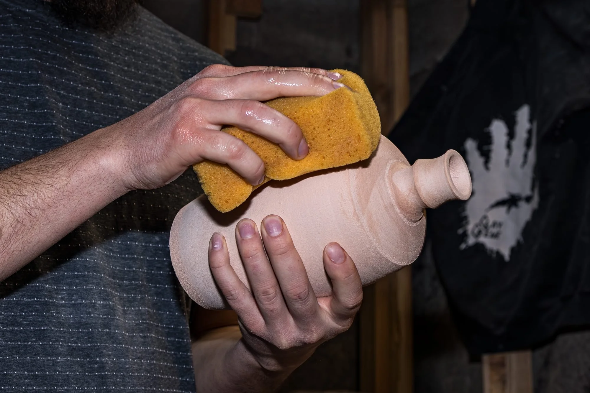 A yellow sponge wiping dust from a bisque fired bottle held in Adam's hands