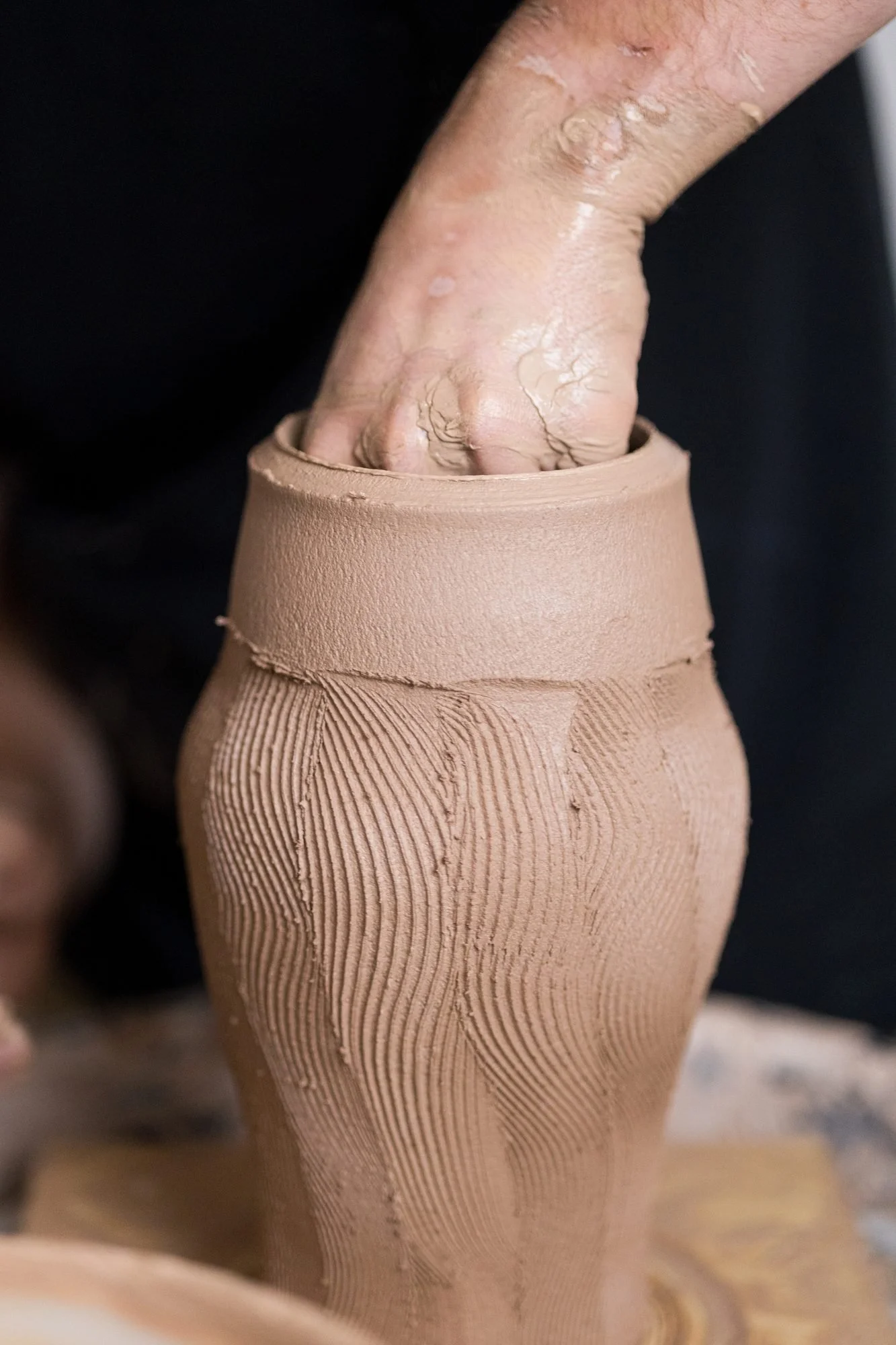 Adam's hand inside a tall textured clay pot as he shapes it on the wheel