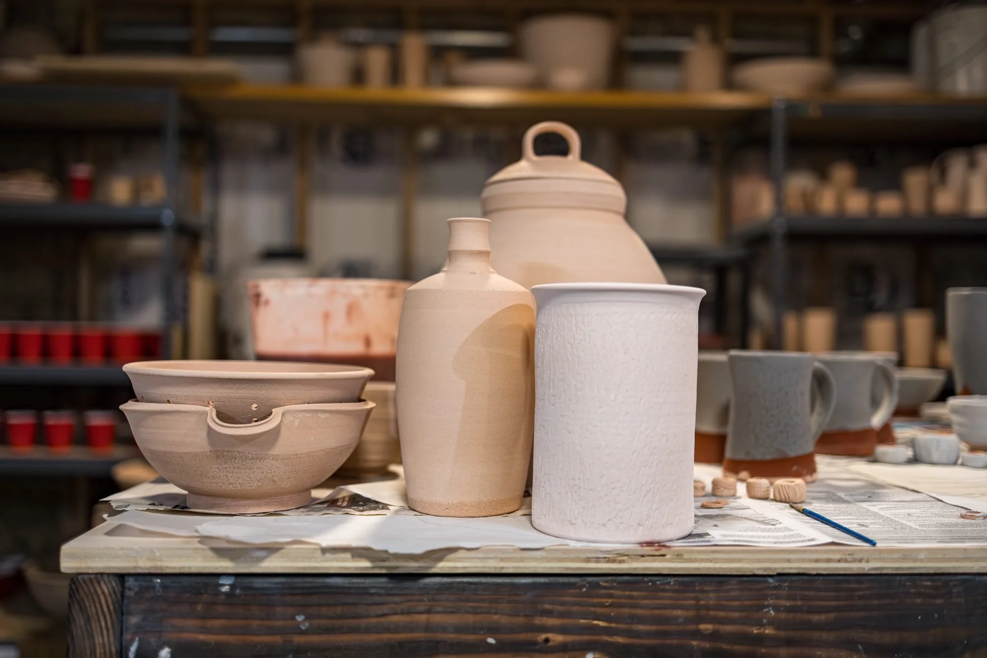 Unfired and bisque pots, jars and bowls grouped on a table in front of studio shelves