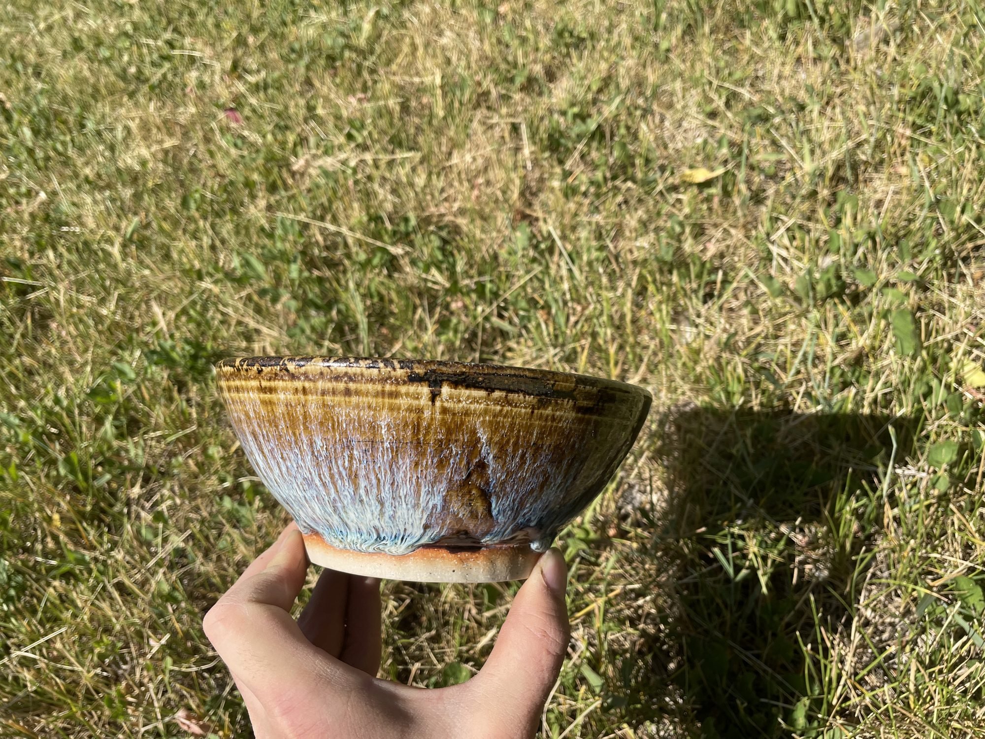 A hand holding a glazed stoneware bowl with brown and blue drips above green grass