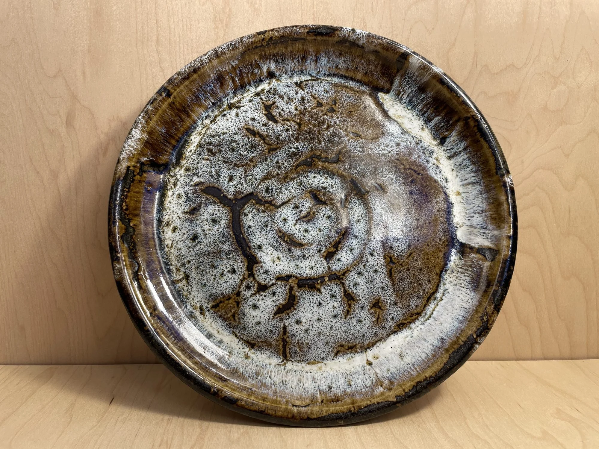 A glazed plate with white glaze breaking over brown, standing upright on a wooden table