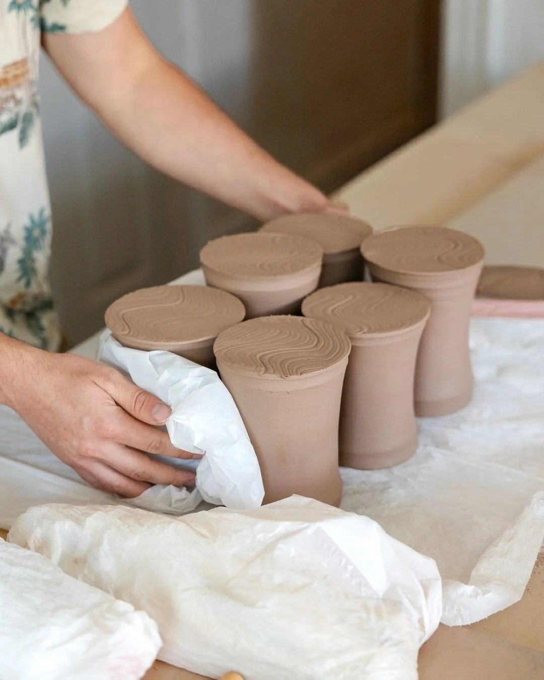These cups are going under plastic.

Clay shrinks as it dries. If the rim dries faster than the base, the top shrinks while the bottom is still soft, the two pull against each other, and the rim splits. Wrapping pots loosely in plastic slows everythi