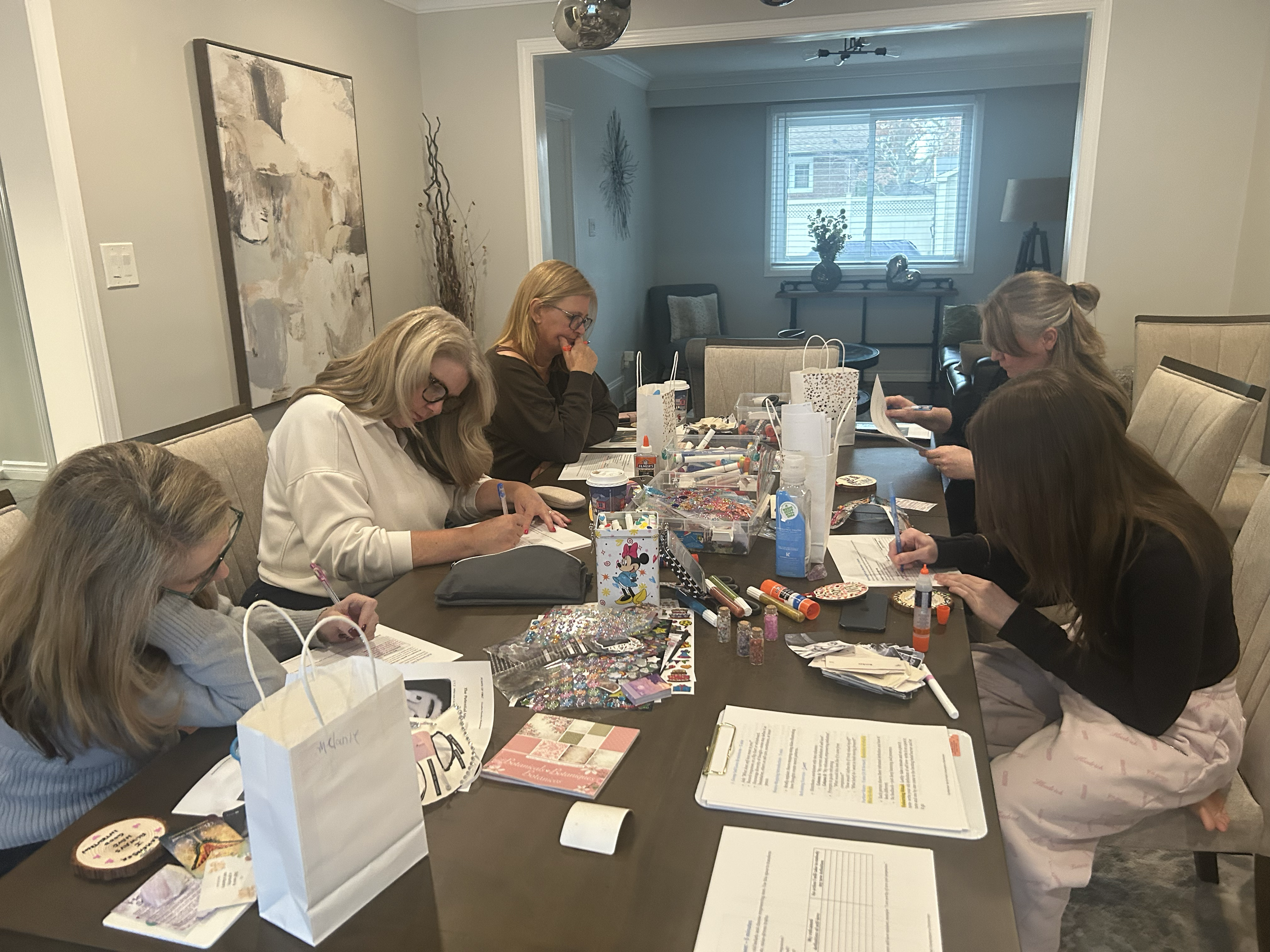 Women sitting around a dining table engaged in arts and crafts, with various craft supplies, papers, and decorative items spread across the table, in a well-lit room with decorated walls and a window in the background.