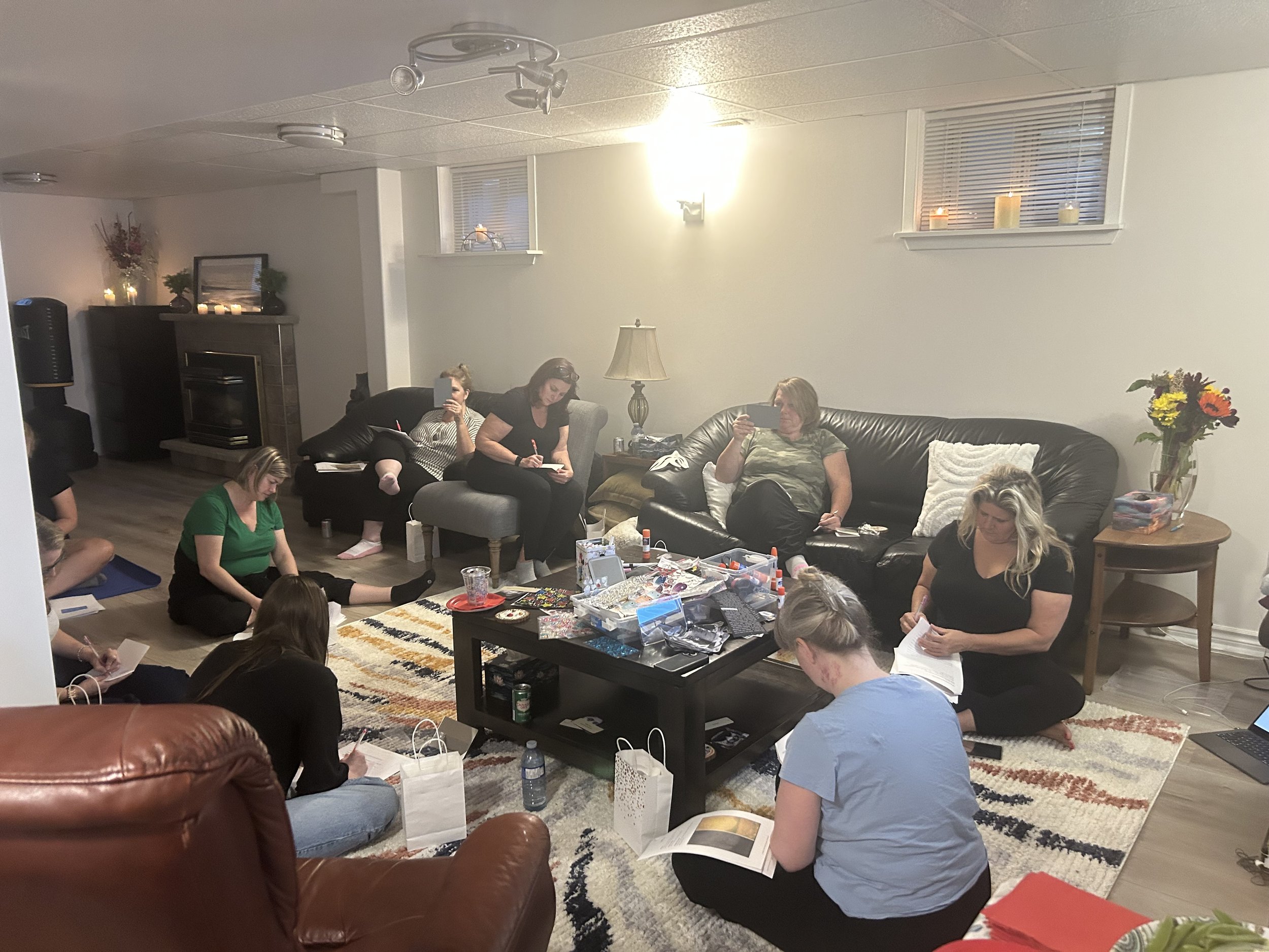 A group of people are sitting and kneeling on a multicolored striped rug in a living room, engaging in a craft or activity. Some are holding papers or notebooks. The room has a dark leather couch with throw pillows, a coffee table with crafting supplies, candles, and flowers. The back wall features a white wall, with two small windows with blinds, lit by a wall sconce and ceiling lights. There is a fireplace with candles and decorations on the mantle.