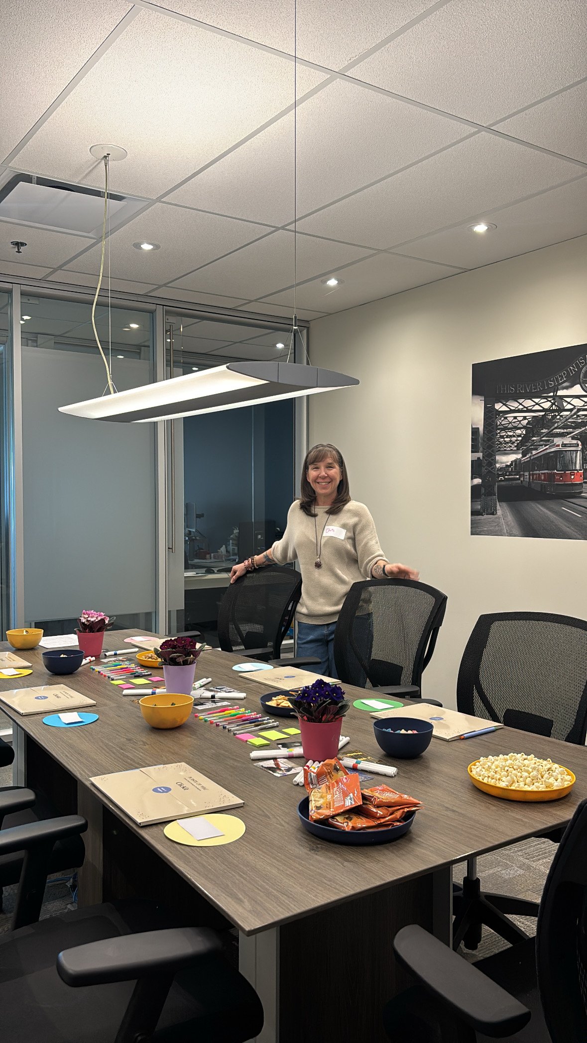 A woman standing in a conference room with a long table, chairs, plants, and snacks, smiling at the camera.