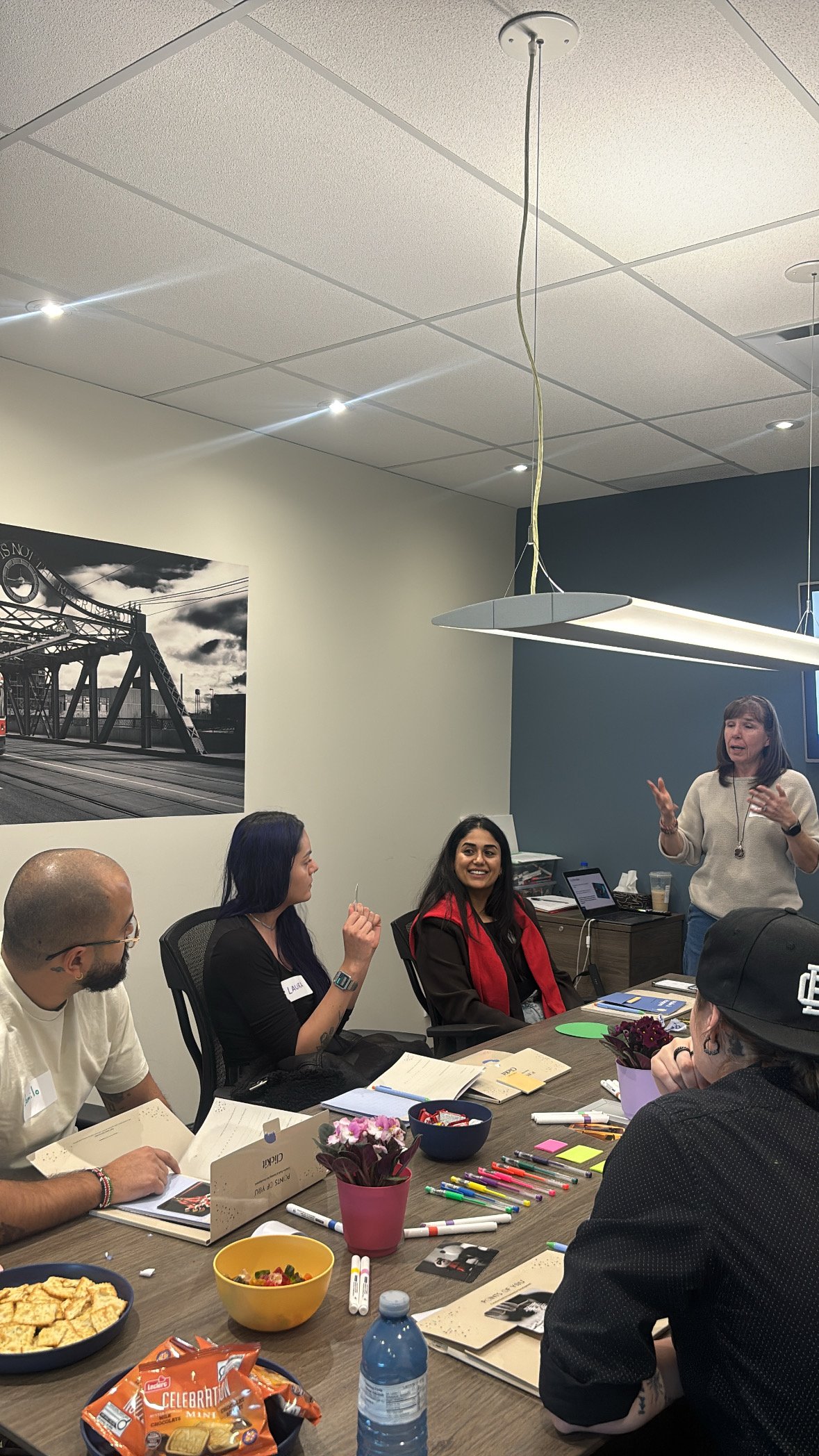 A group of people in a meeting room are engaged in a discussion, with snacks and supplies on the table. A woman stands and speaks while others listen and look at her.