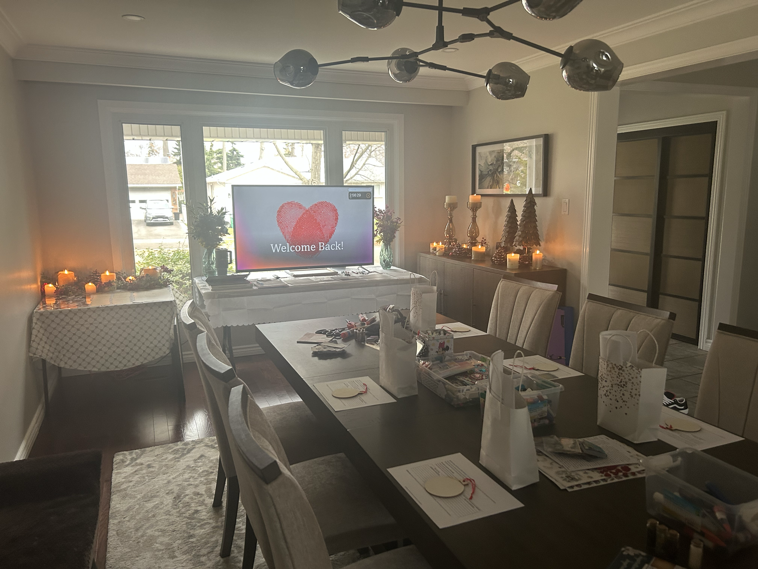 Interior of a living room with a dining table, chairs, a sideboard decorated with candles and small Christmas trees, and a television with a welcome back message displaying a red heart in the background.