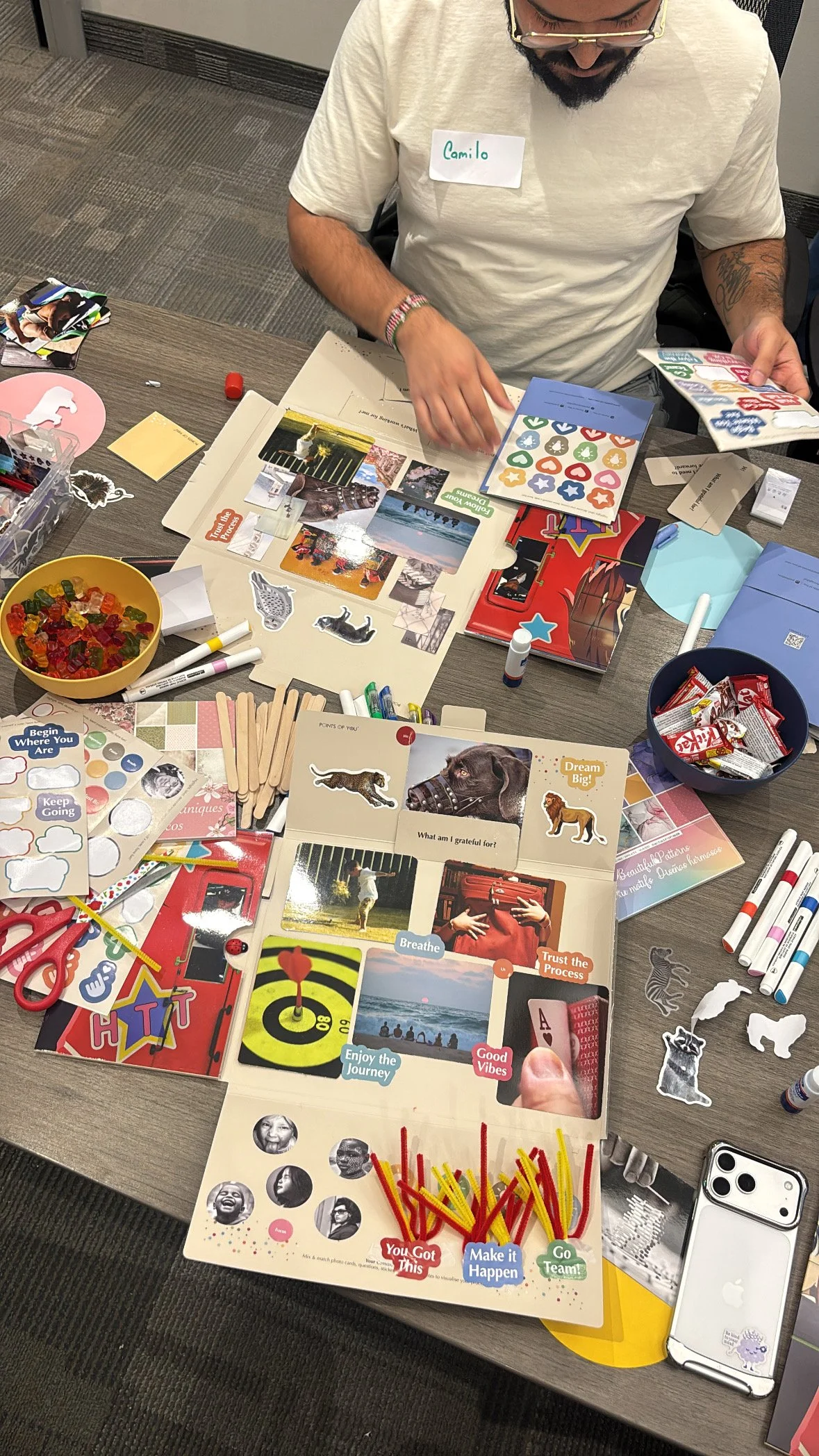 A person with a name tag labeled 'Camilo' sitting at a table filled with various craft supplies, stickers, photos, markers, and paper for a creative activity or project.