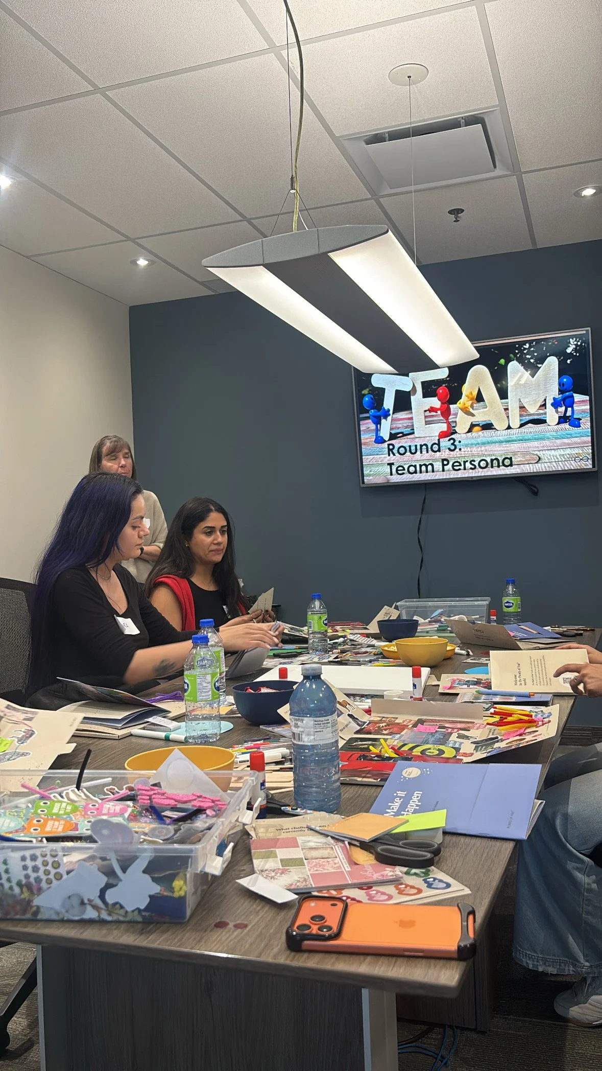 People sitting around a table in a meeting room with craft supplies and water bottles, watching a presentation on a wall-mounted screen that says 'Team' and 'Round 3: Team Persona'.