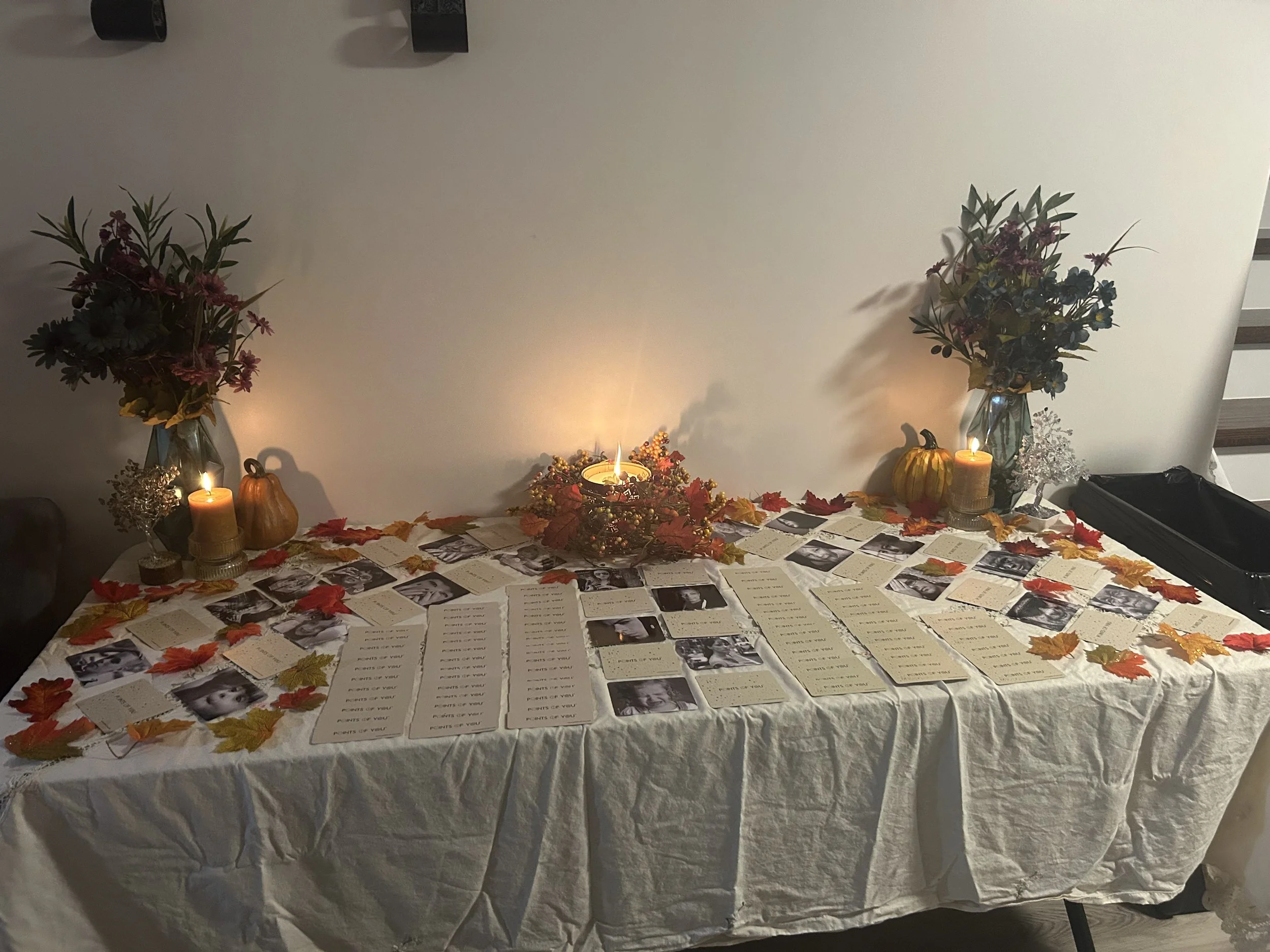 A table covered with a white tablecloth displays black and white photographs, handwritten notes, and autumn leaves. Two large flower arrangements in vases and lit candles decorate the background.