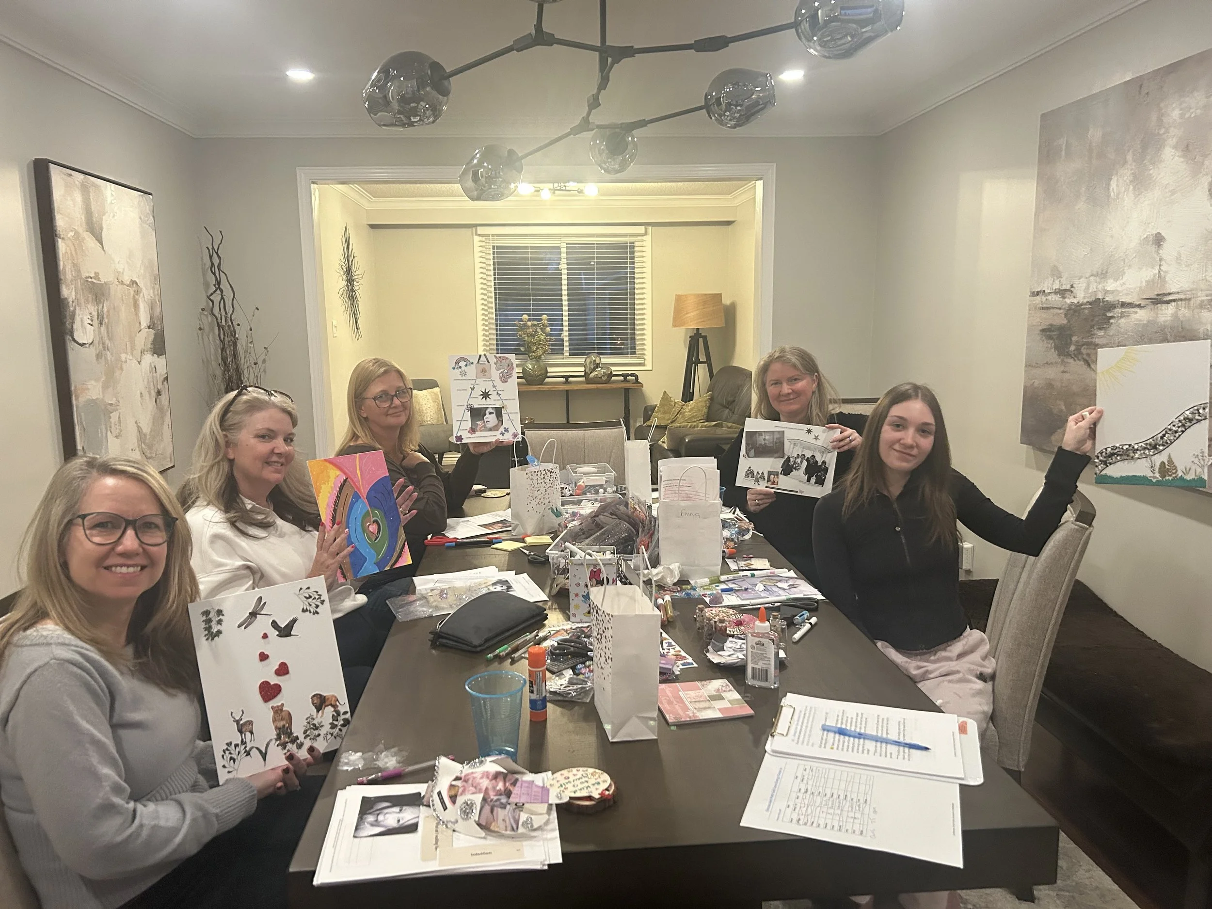 A group of women sitting at a table, holding various finished artwork projects, in a room with modern decorations and large artwork on the walls, engaged in arts and crafts activities.