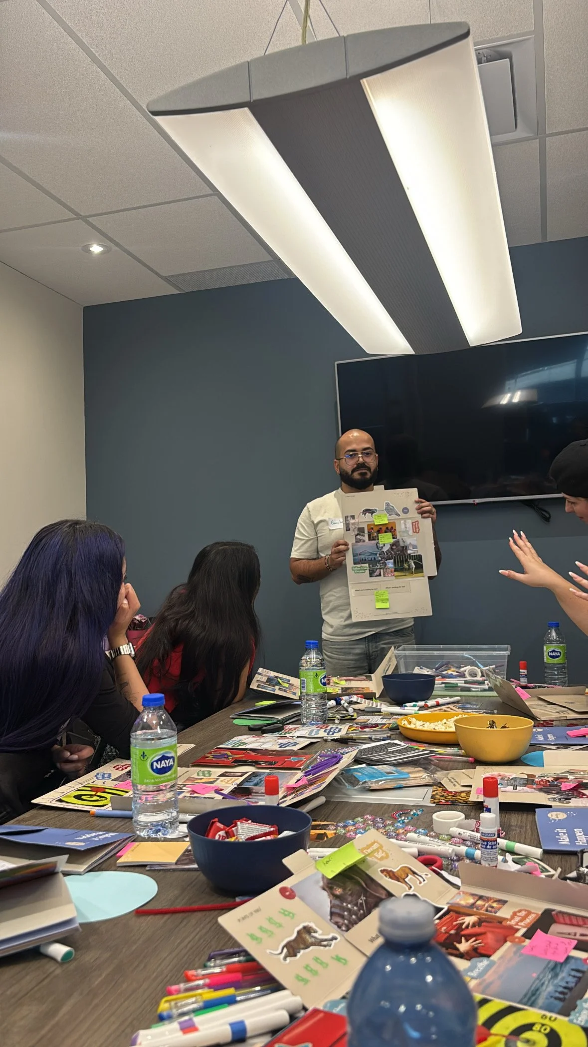 A man standing and presenting a collage to an audience in a room with stained and a black wall, with table cluttered with art supplies, snacks, and water bottles, and people seated and engaged.