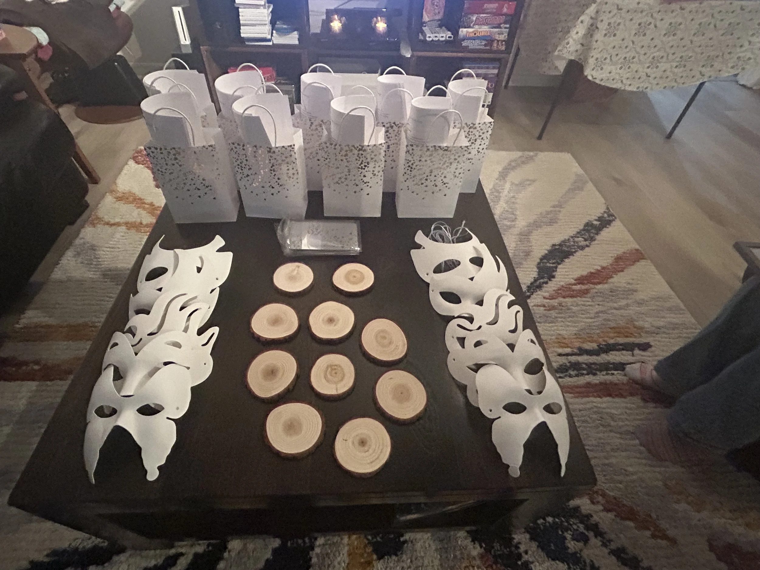 Table with multiple white and clear gift bags, small wooden logs, and white masks with holes, in a room with a rainbow-colored rug and a bookshelf in the background.