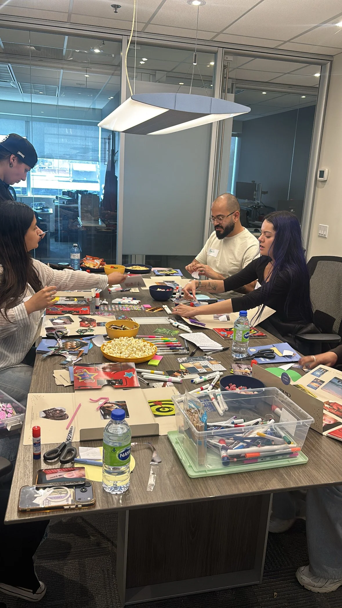 People sitting around a conference table engaged in arts and crafts activities with markers, photographs, scissors, and snacks in a modern office setting.
