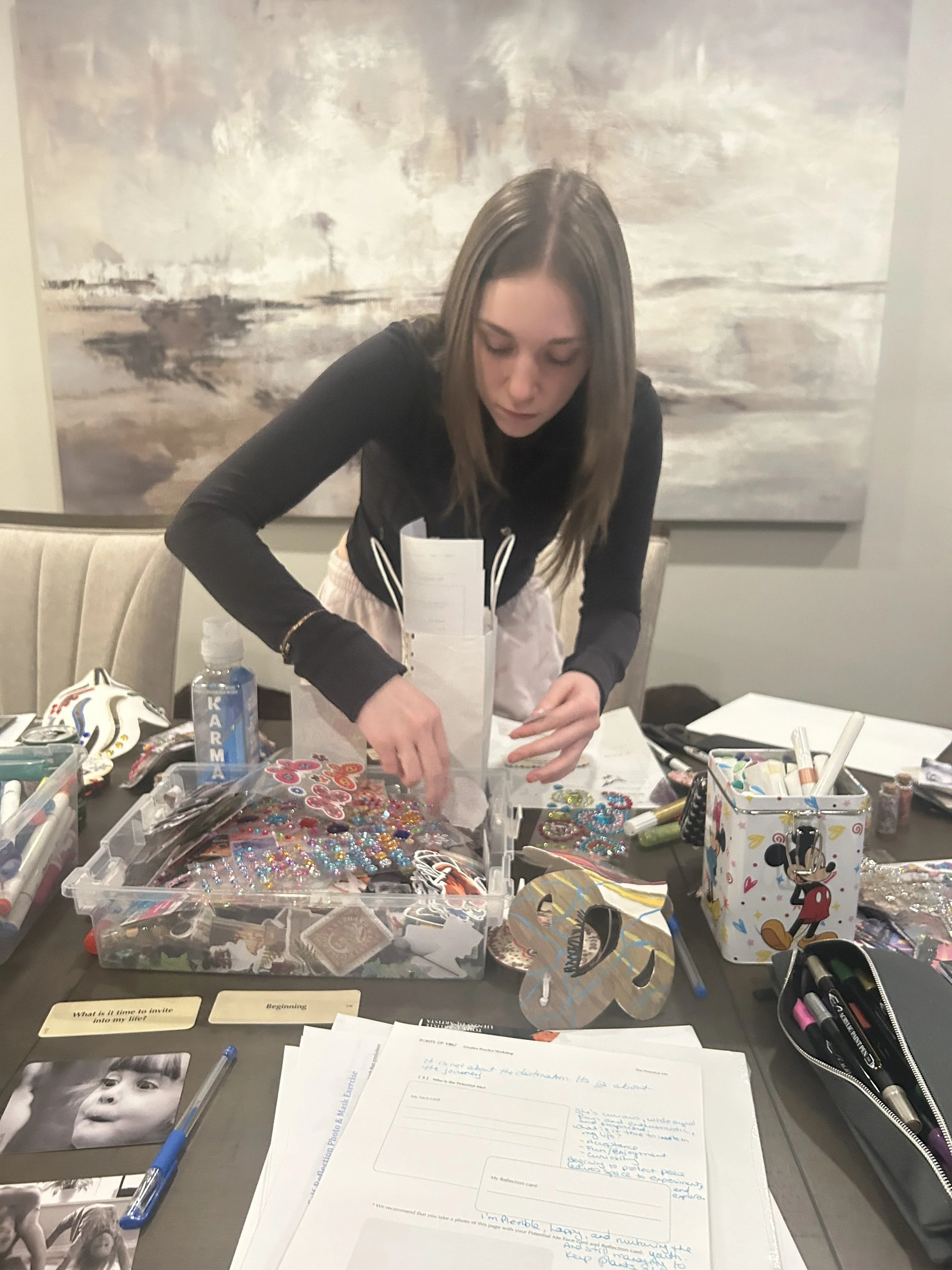 Girl sitting at a table, organizing colorful beads and craft supplies, with papers, markers, a photo, and a Mickey Mouse-themed cup on the table, and an abstract painting on the wall behind her.
