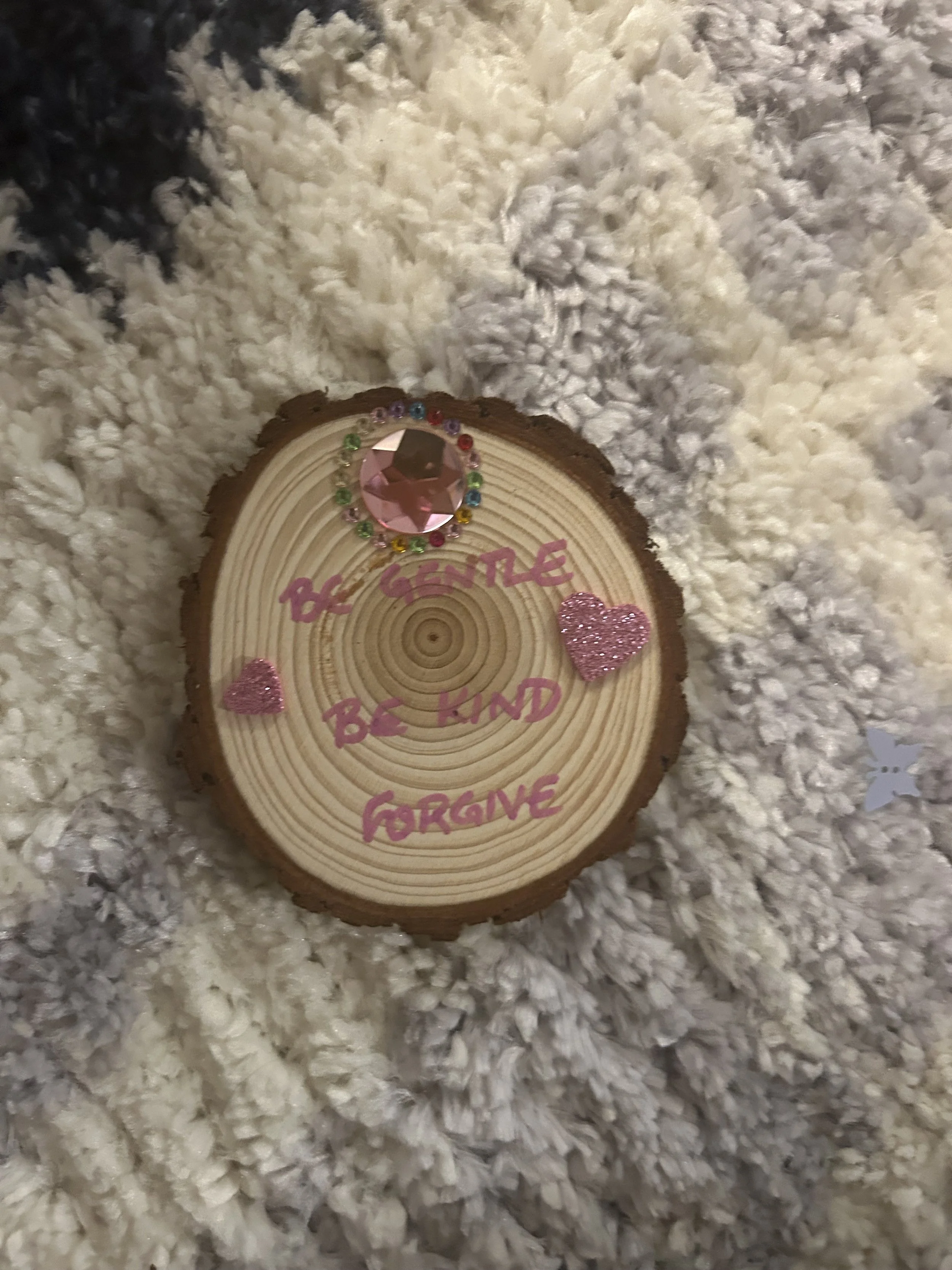 A wooden log slice decorated with pink and multicolored rhinestones, pink glitter hearts, and the handwritten message 'Be Gentle, Be Kind, Be Forgiving'. It is placed on a fluffy white textured surface.